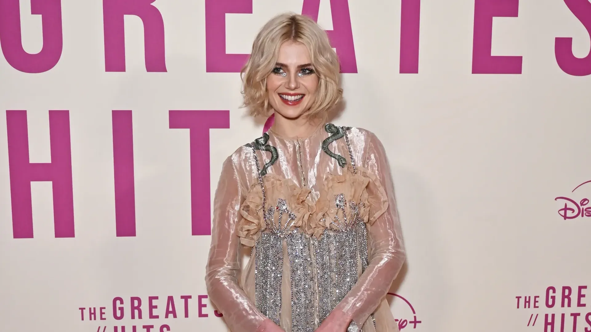 A girl with a short haircut and light hair is standing by the photo wall at the film premiere