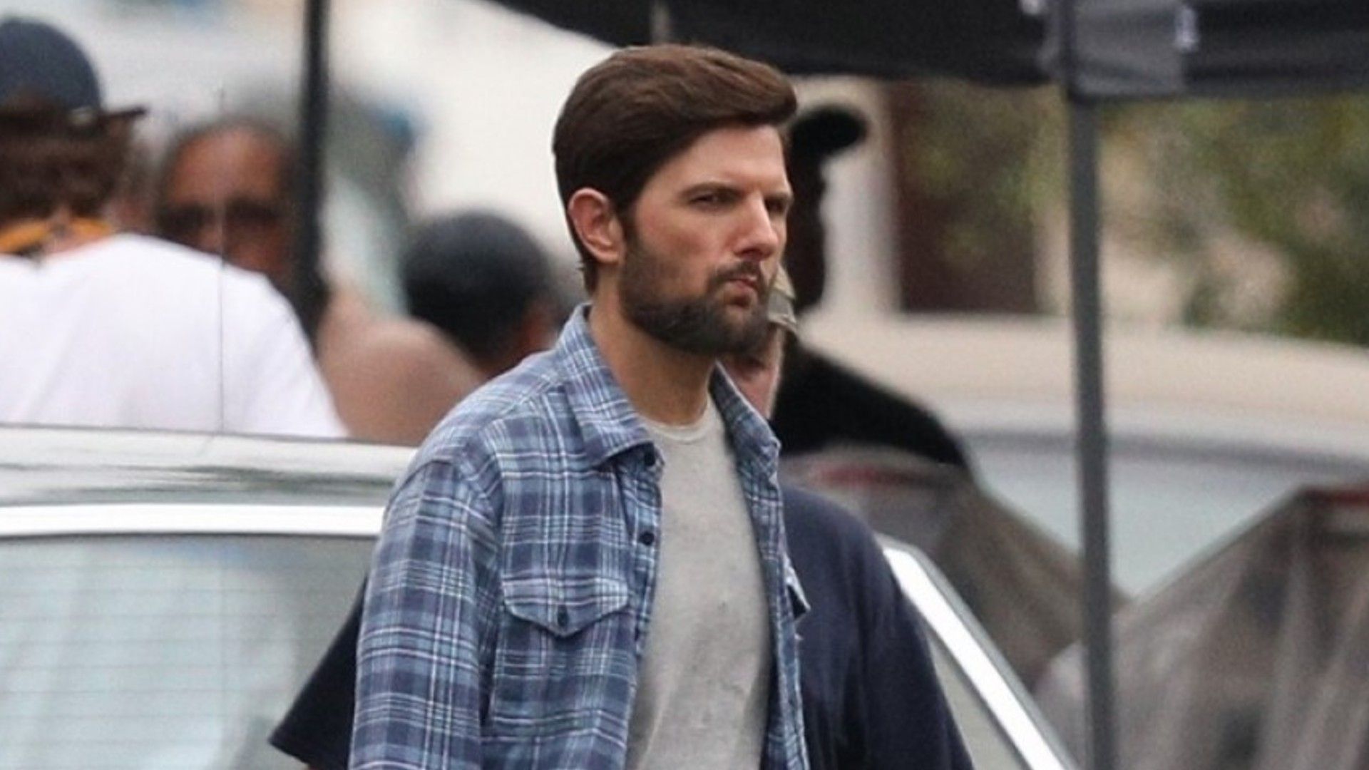 A brown-haired man in a checkered shirt walks through the city, relaxed posture