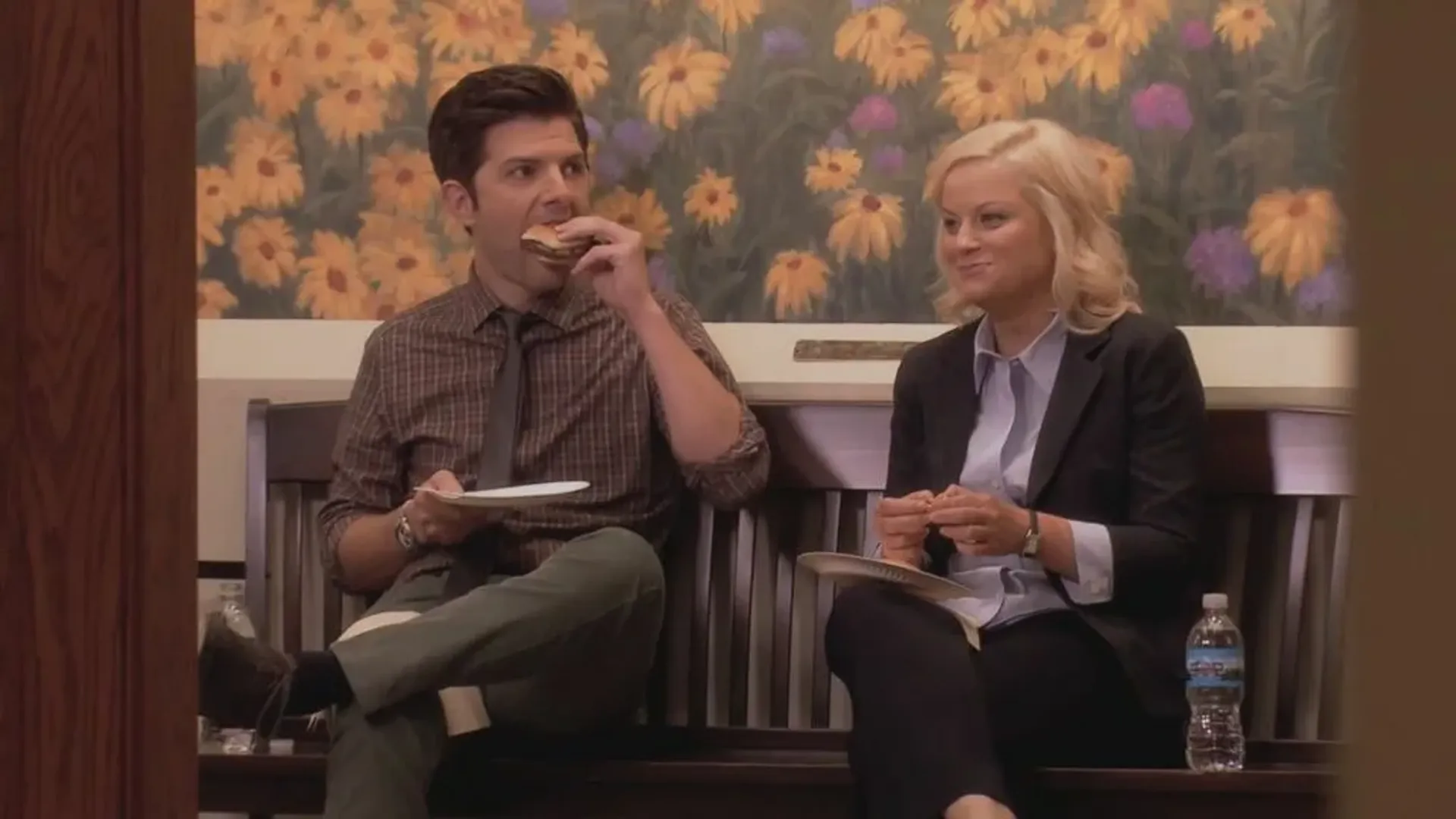 A man and woman sitting on chairs in the hallway, the man is eating a hamburger