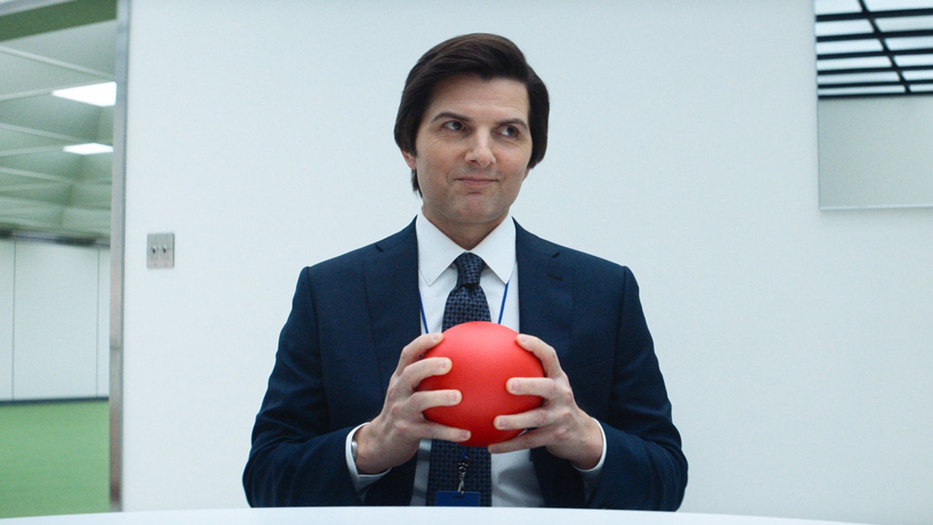 A middle-aged man in a suit holds a red ball in his hands, with white walls in the background