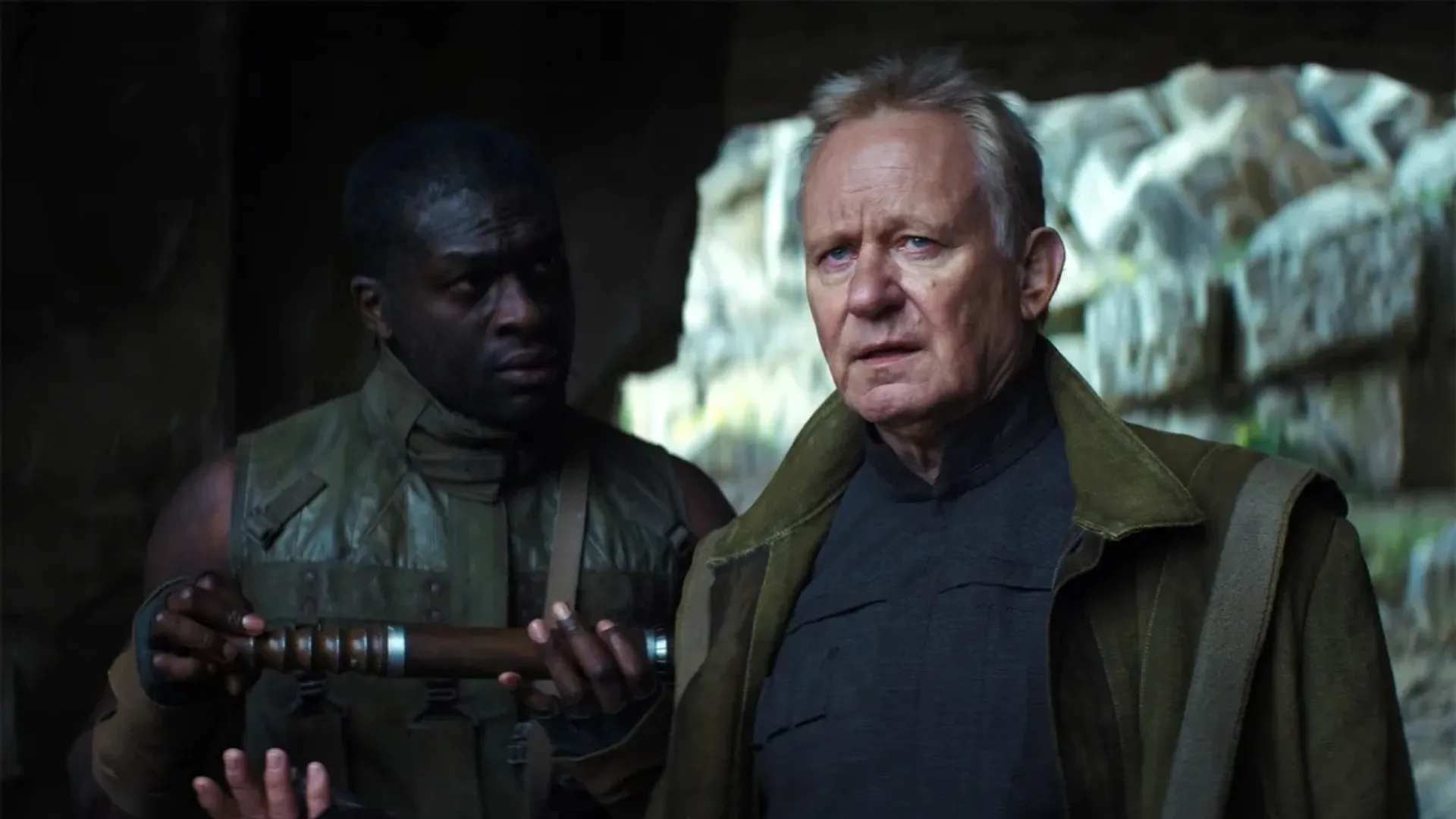 An elderly man in a uniform resembling military attire stands in a cave, being addressed by a man holding a telescope