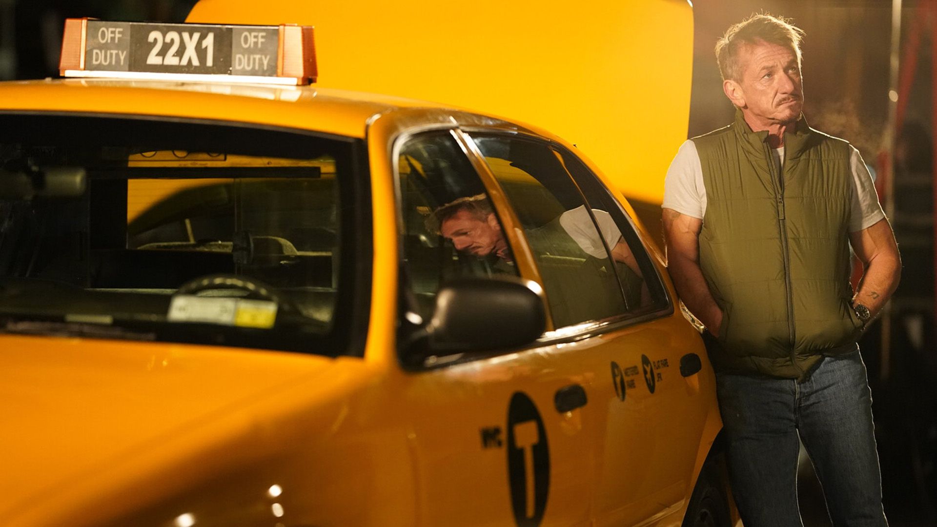 Sean Penn in a green vest leaning against a yellow taxi cab