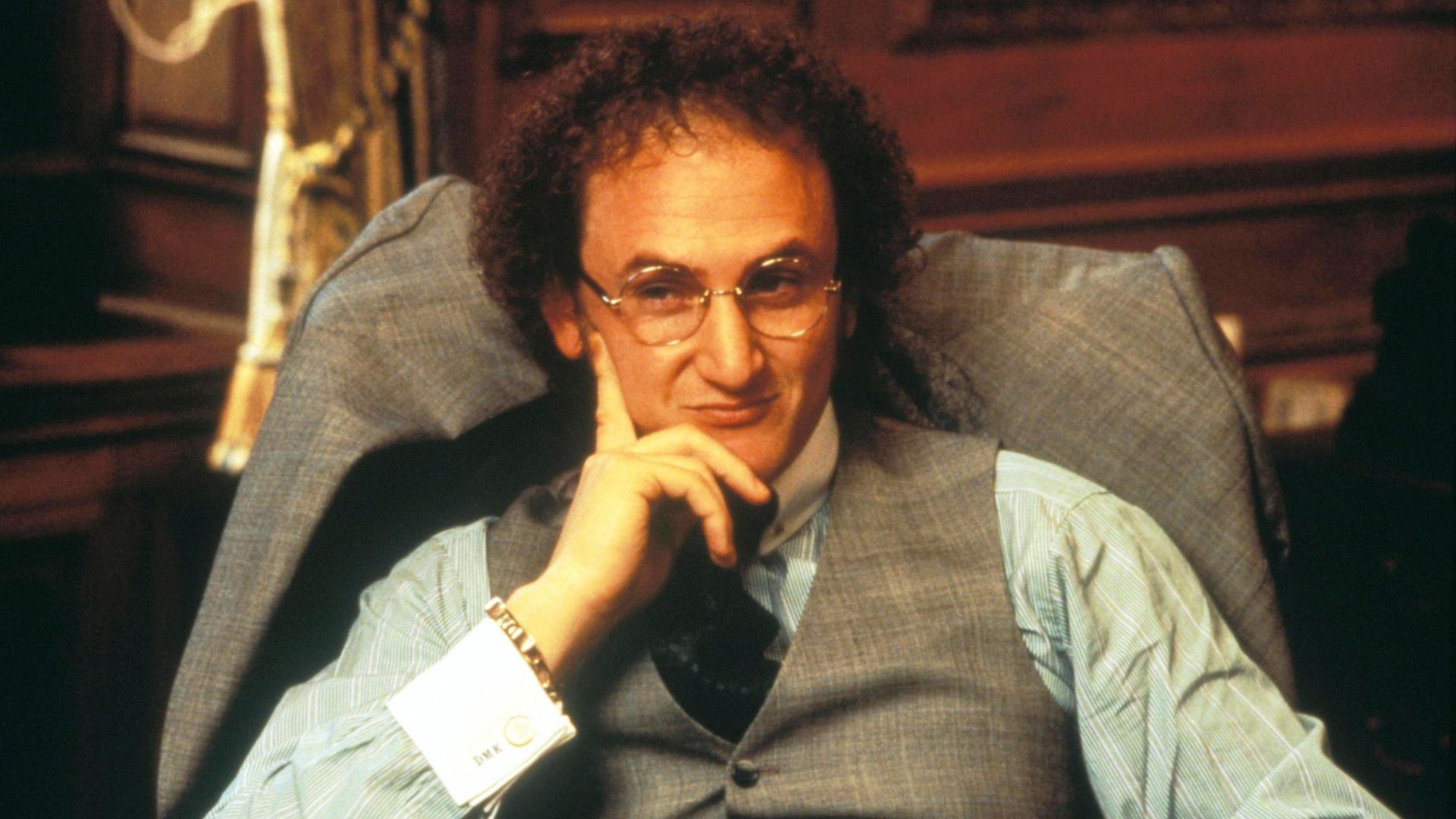 Sean Penn with curly hair and round glasses sitting in a chair