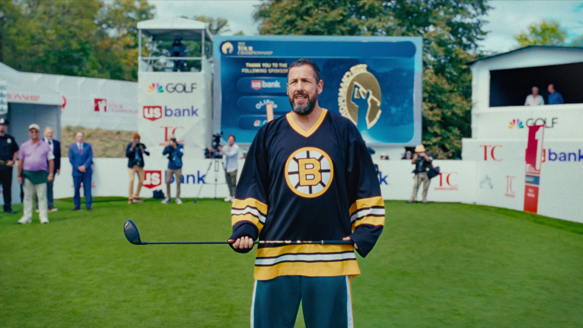 Adam Sandler in a jersey holds a hockey stick while standing in a stadium