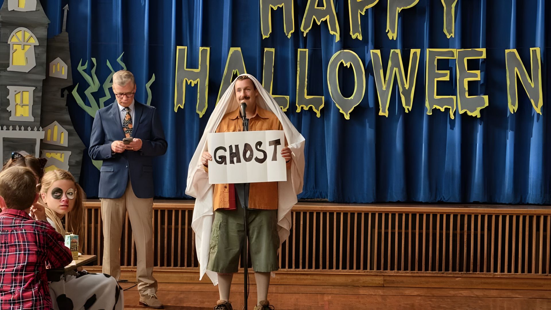 Adam Sandler wearing a bedsheet over his head, holding a sign that says GHOST