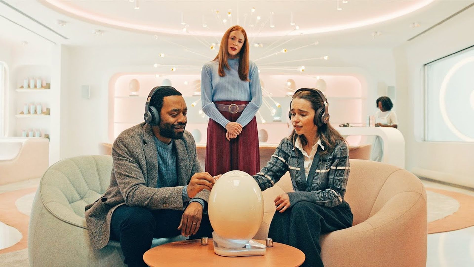 Emilia Clarke and Chiwetel Ejiofor sitting in chairs, futuristic setting, scene from The Pod Generation