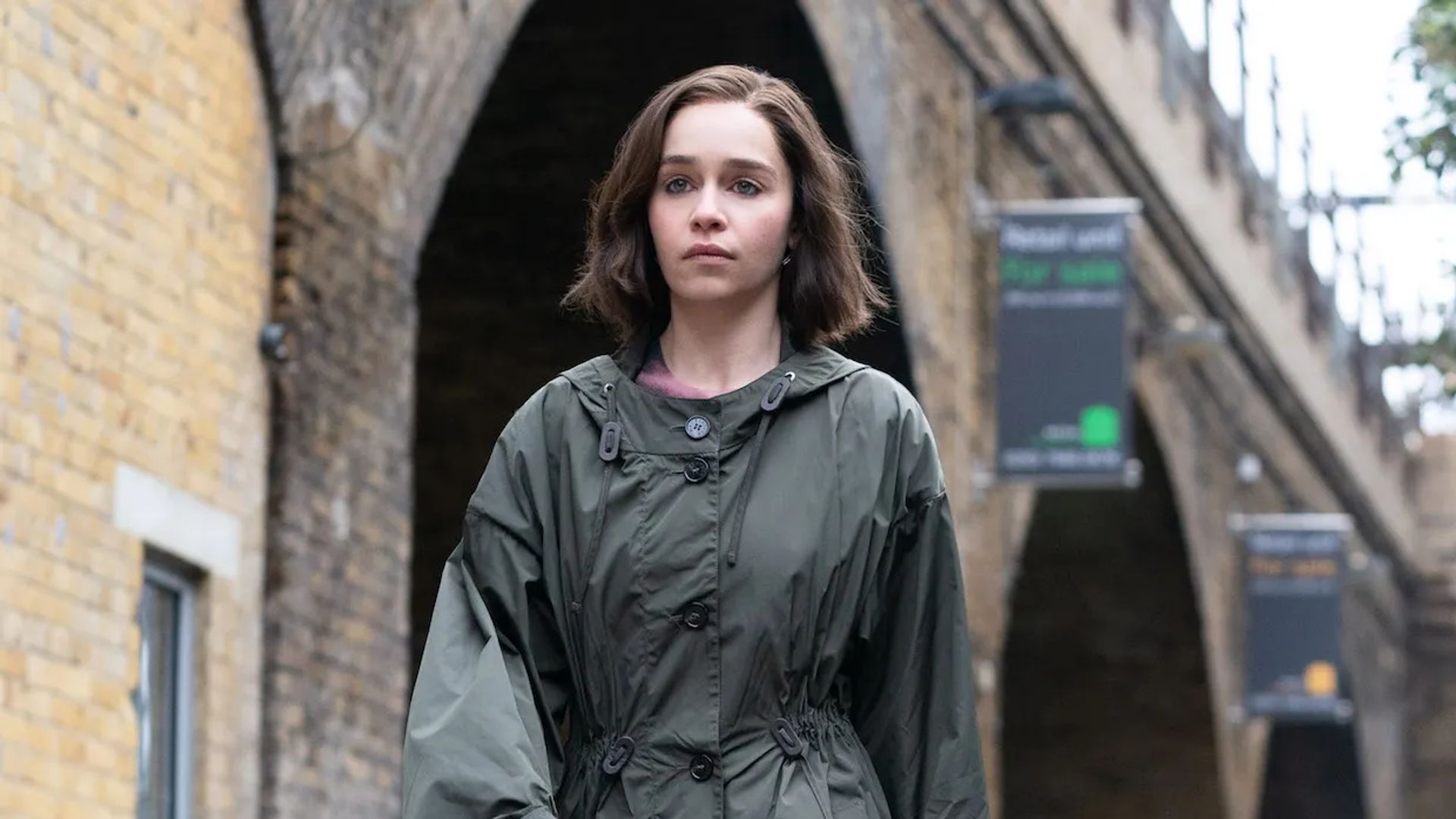 Emilia Clarke in a raincoat walking down the street, focused gaze ahead