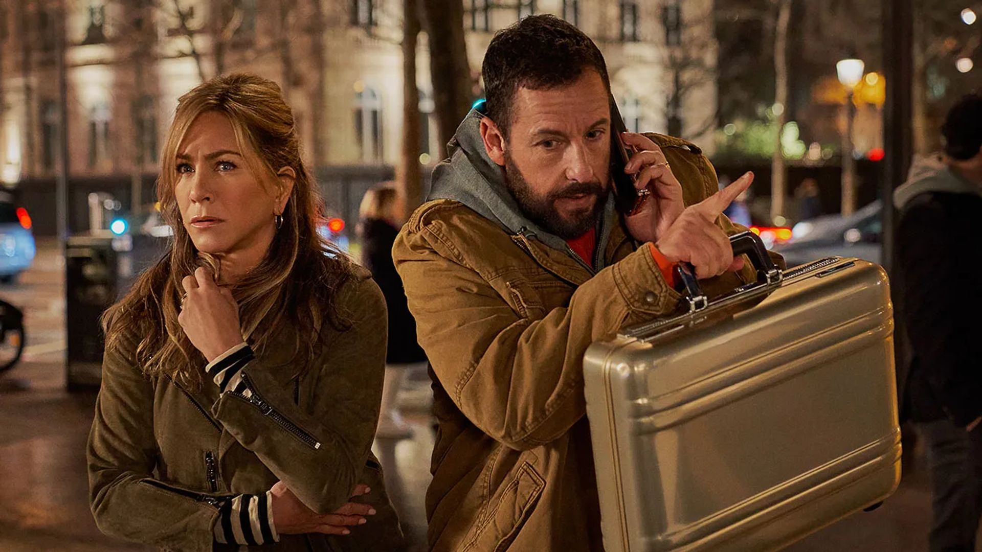 Jennifer Aniston and Adam Sandler on a street, she looks frightened, he's talking on the phone and holding a briefcase