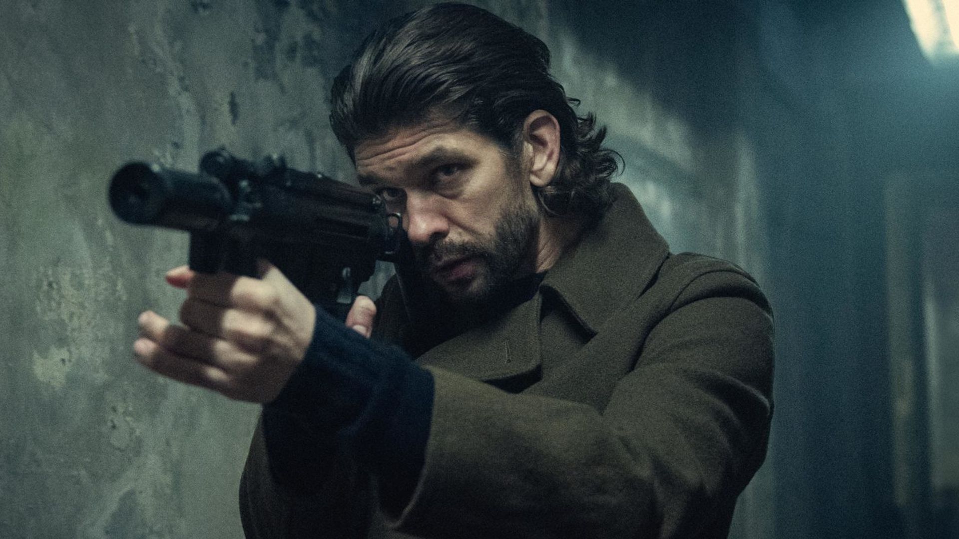 Ben Whishaw with long hair in a coat holding a gun