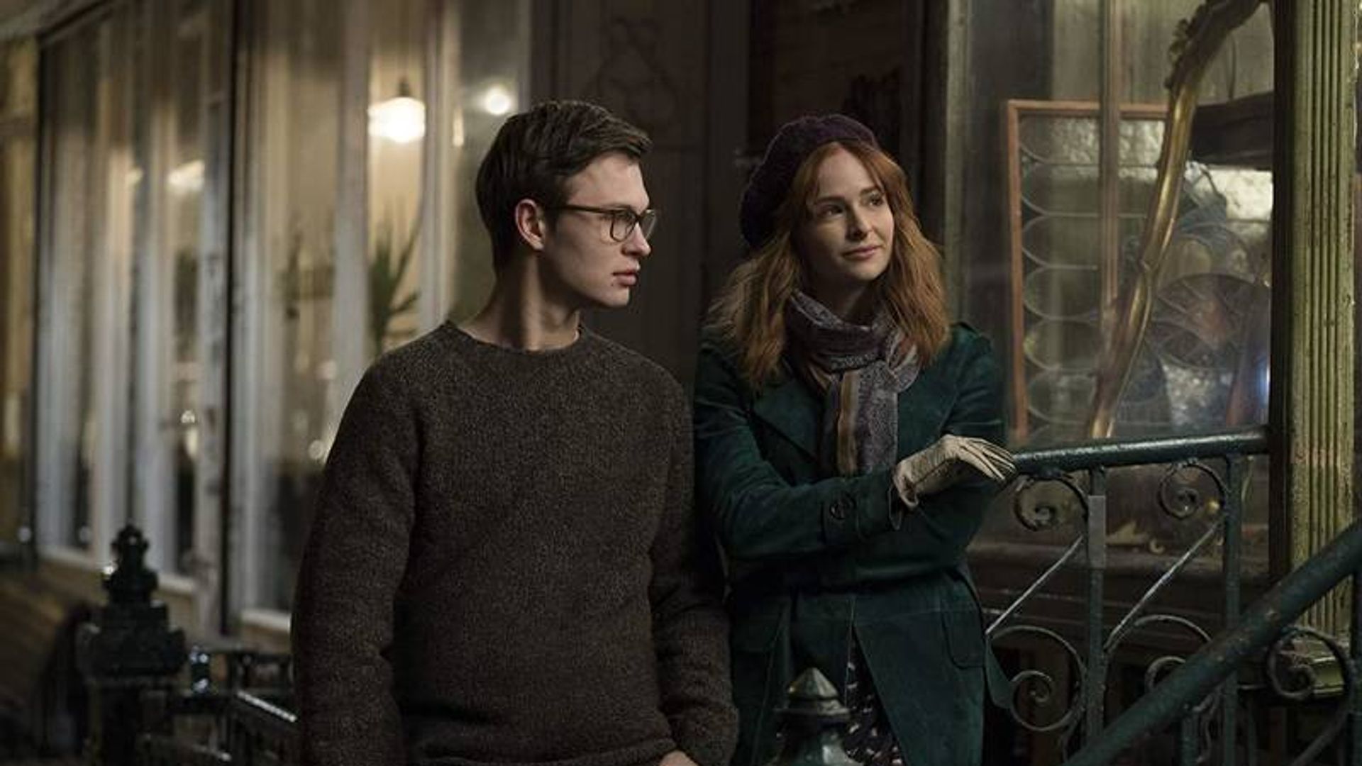 A young man wearing glasses and a woman in a coat and beret talking while standing by a railing on the street in the evening