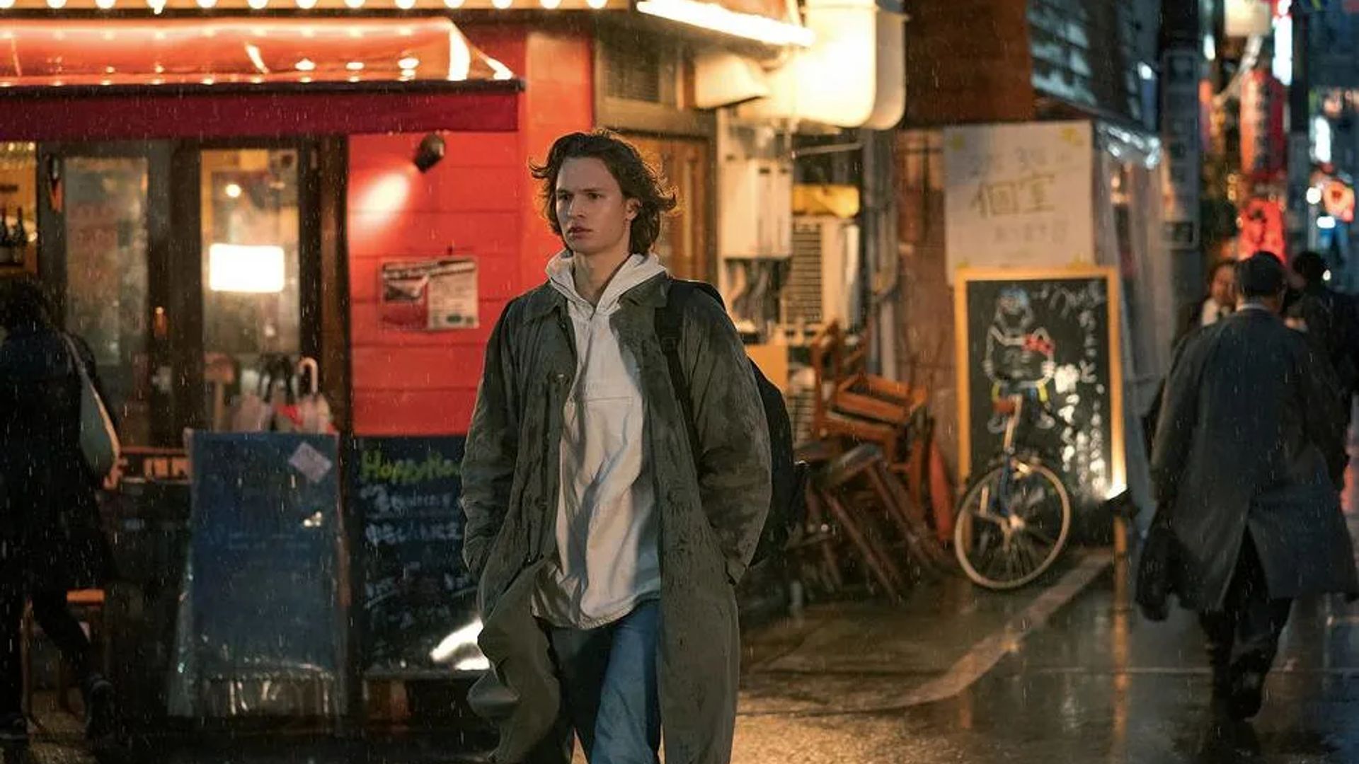 Ansel Elgort in a coat and hoodie walks down a rainy street past brightly lit storefronts and restaurants