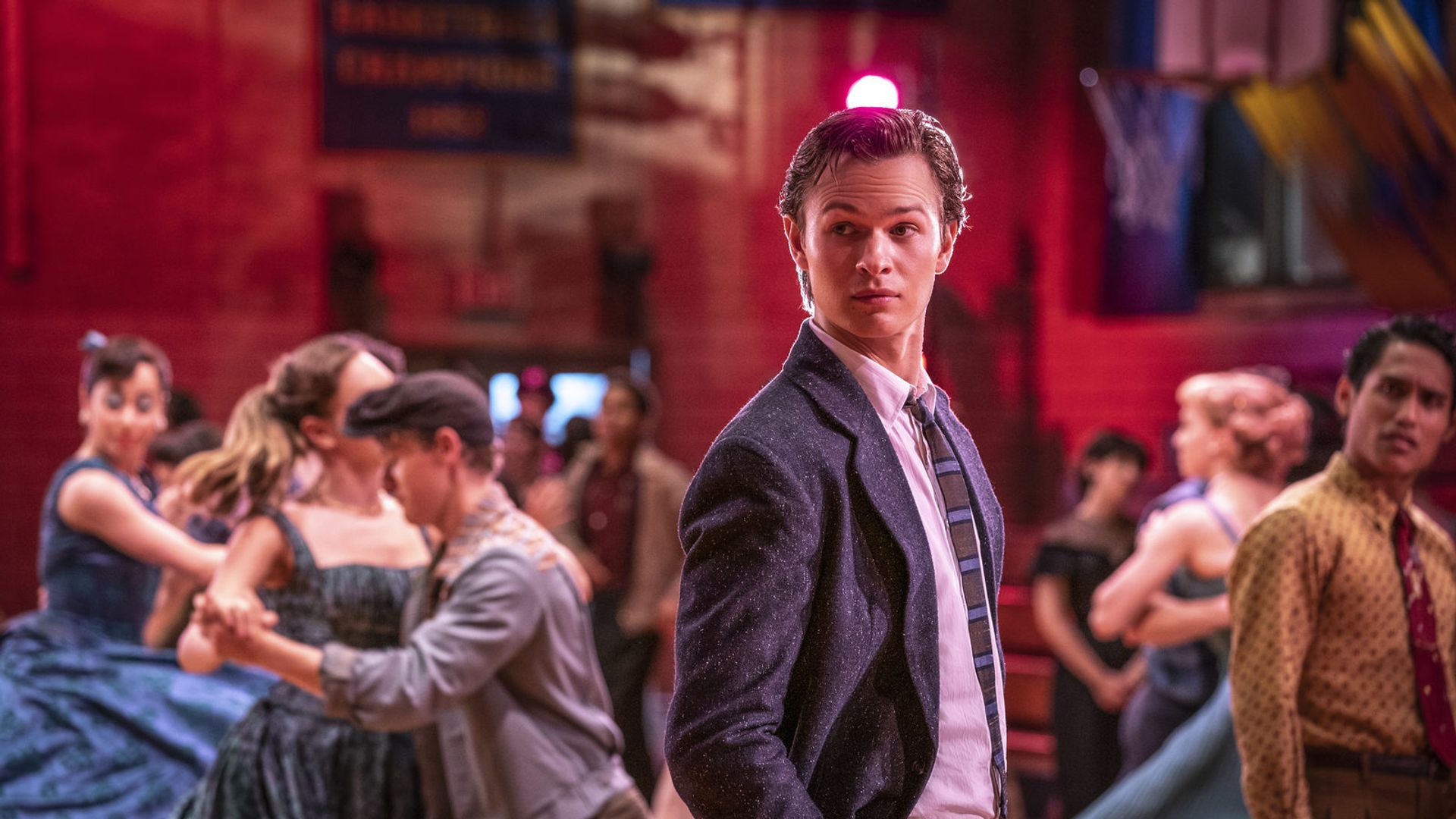 Ansel Elgort in a suit and tie stands at a school dance with couples swirling around him