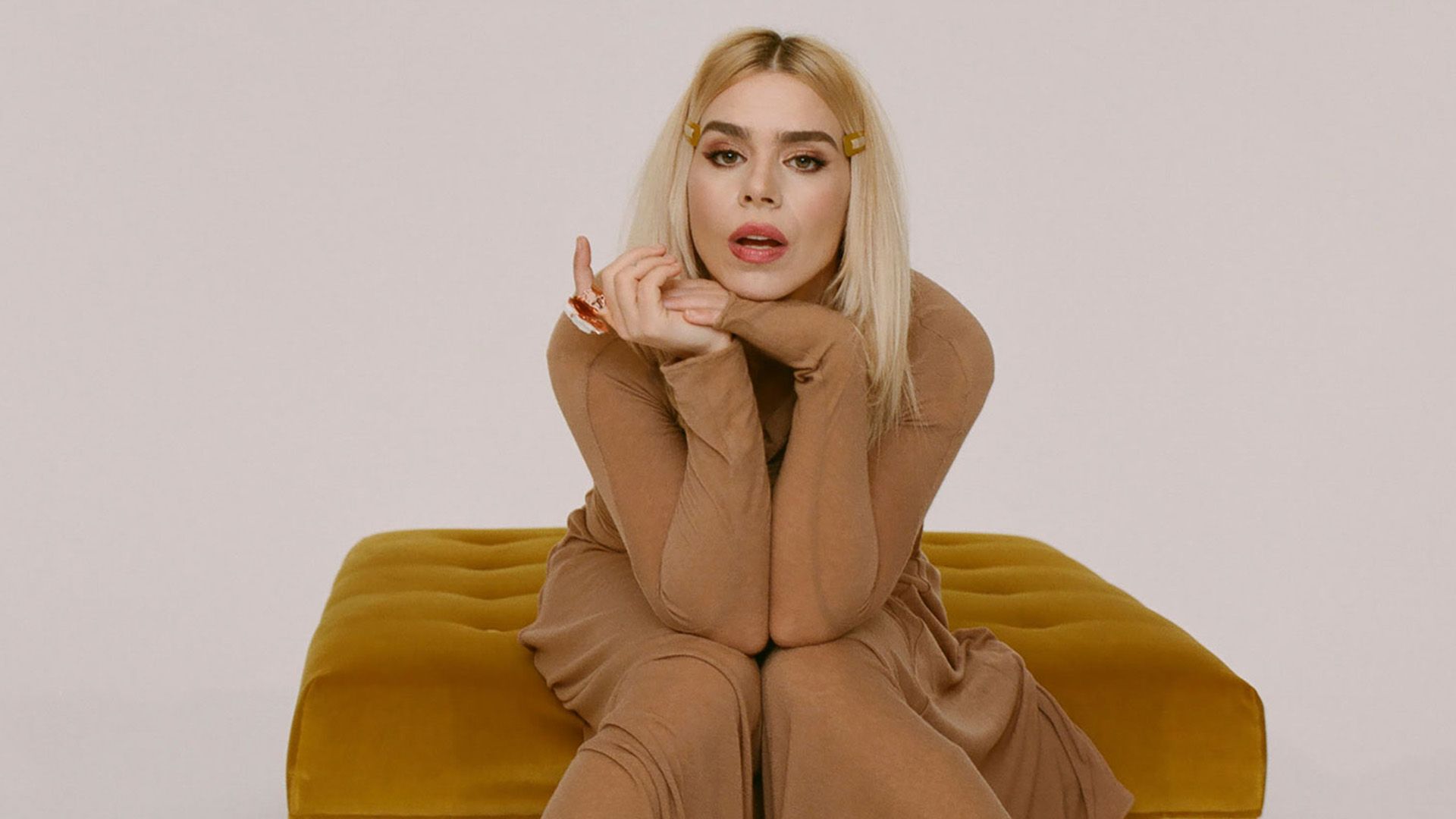 Billie Piper in a long beige dress sits in a ginger armchair