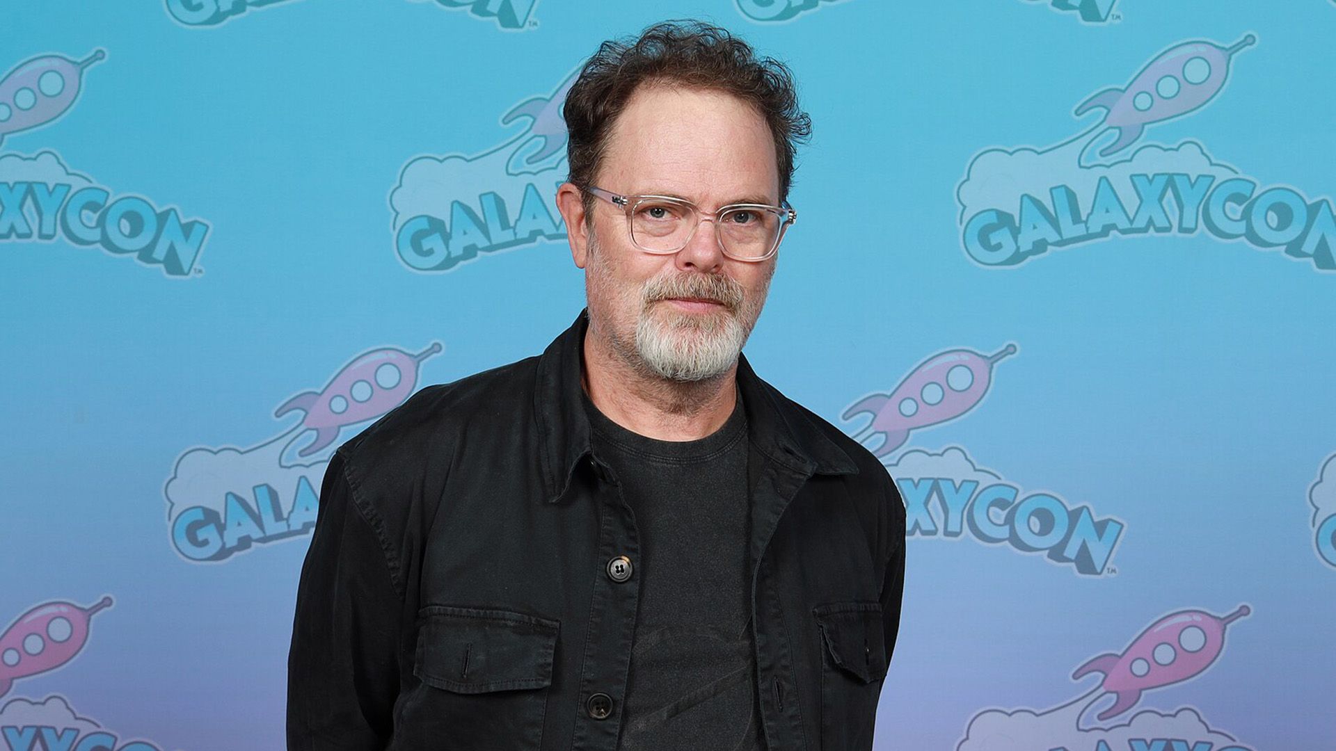 Actor Rainn Wilson wearing glasses and a black jacket against a blue background