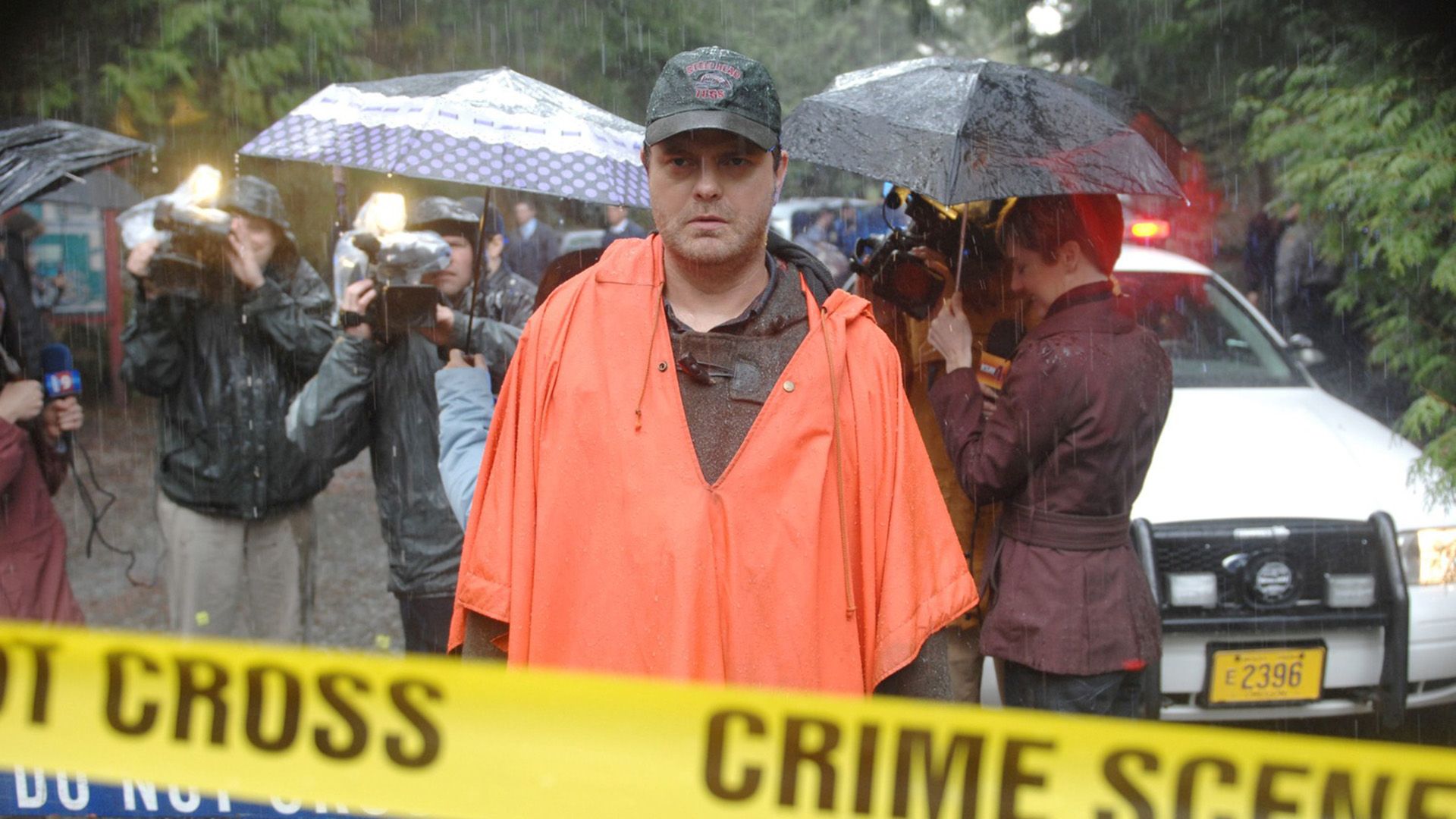Rainn Wilson in an orange raincoat standing in the rain behind yellow police tape
