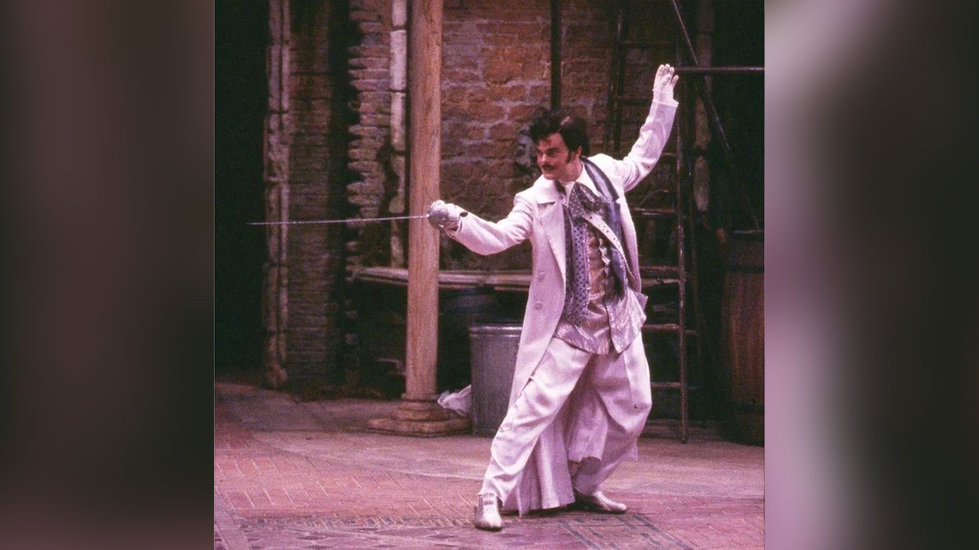 Rainn Wilson in white pants and a white cloak stands holding a sword