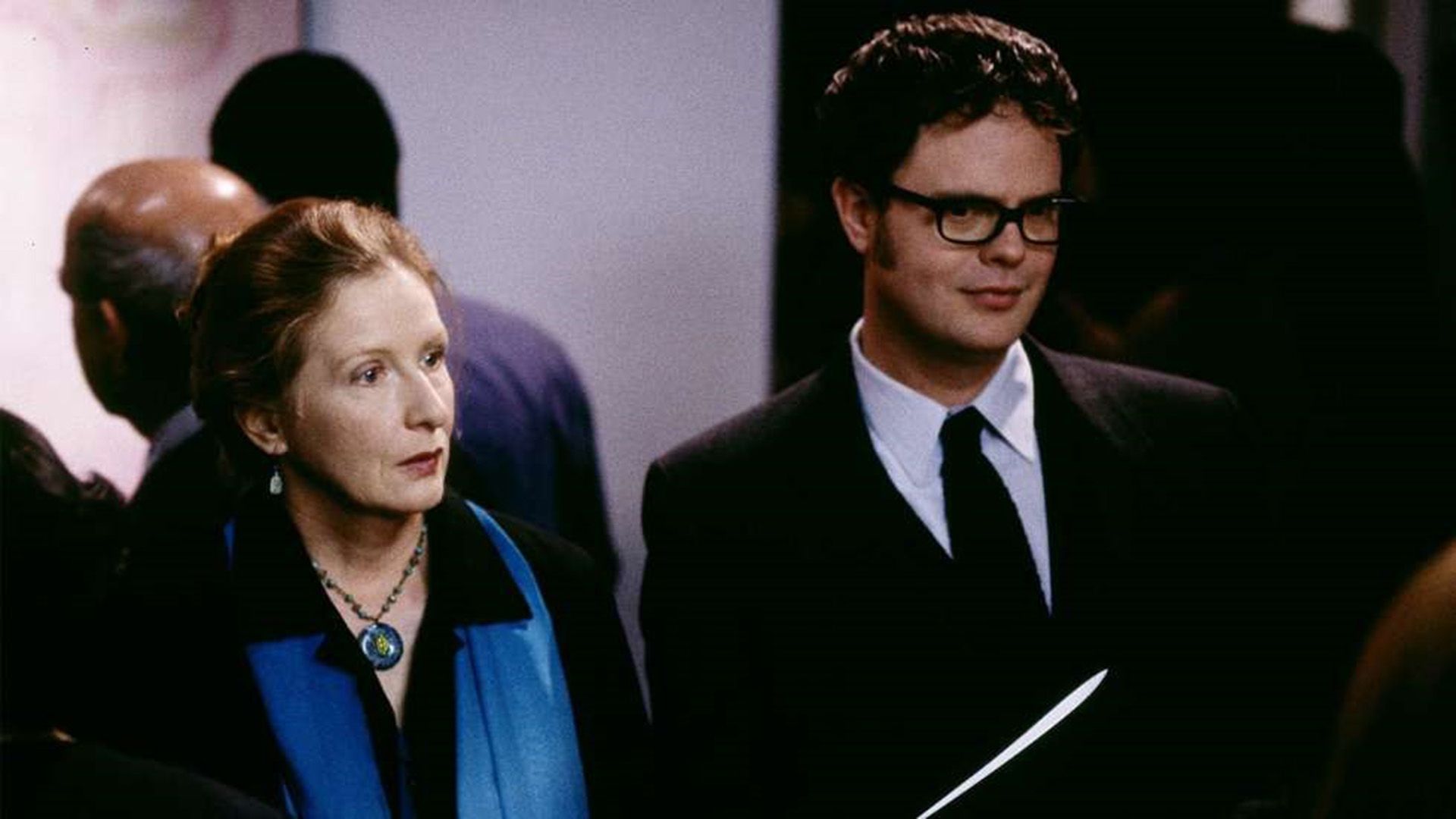 Rainn Wilson wearing glasses and a blazer, standing next to Frances Conroy in a blue scarf