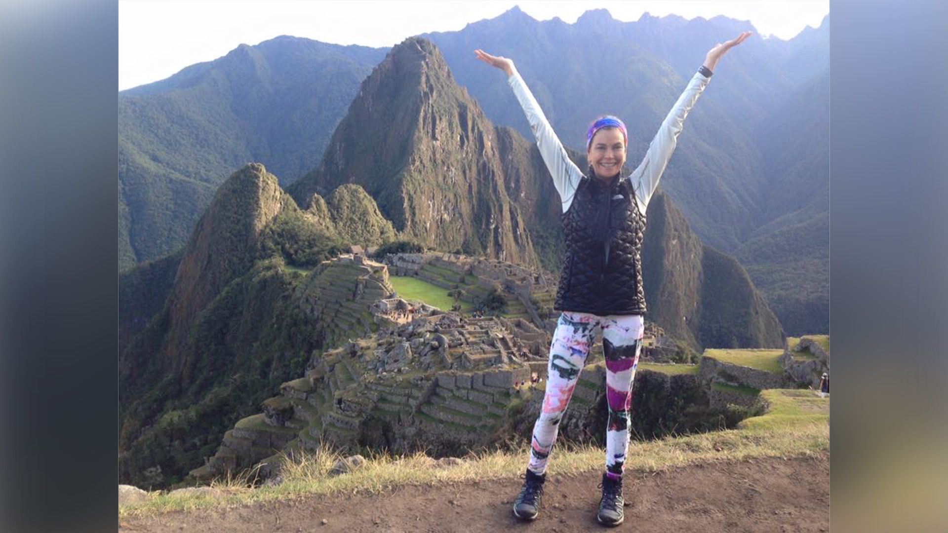 Teri Hatcher in athletic wear stands atop a mountain with her arms raised