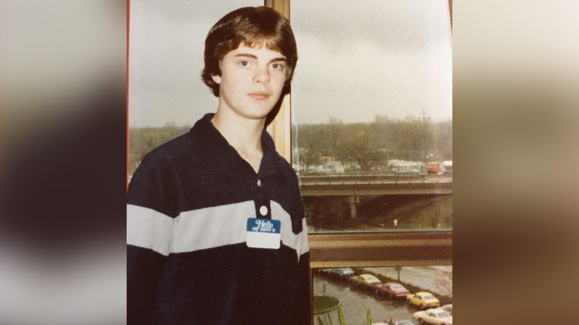 Young Rainn Wilson in a blue polo standing by the window