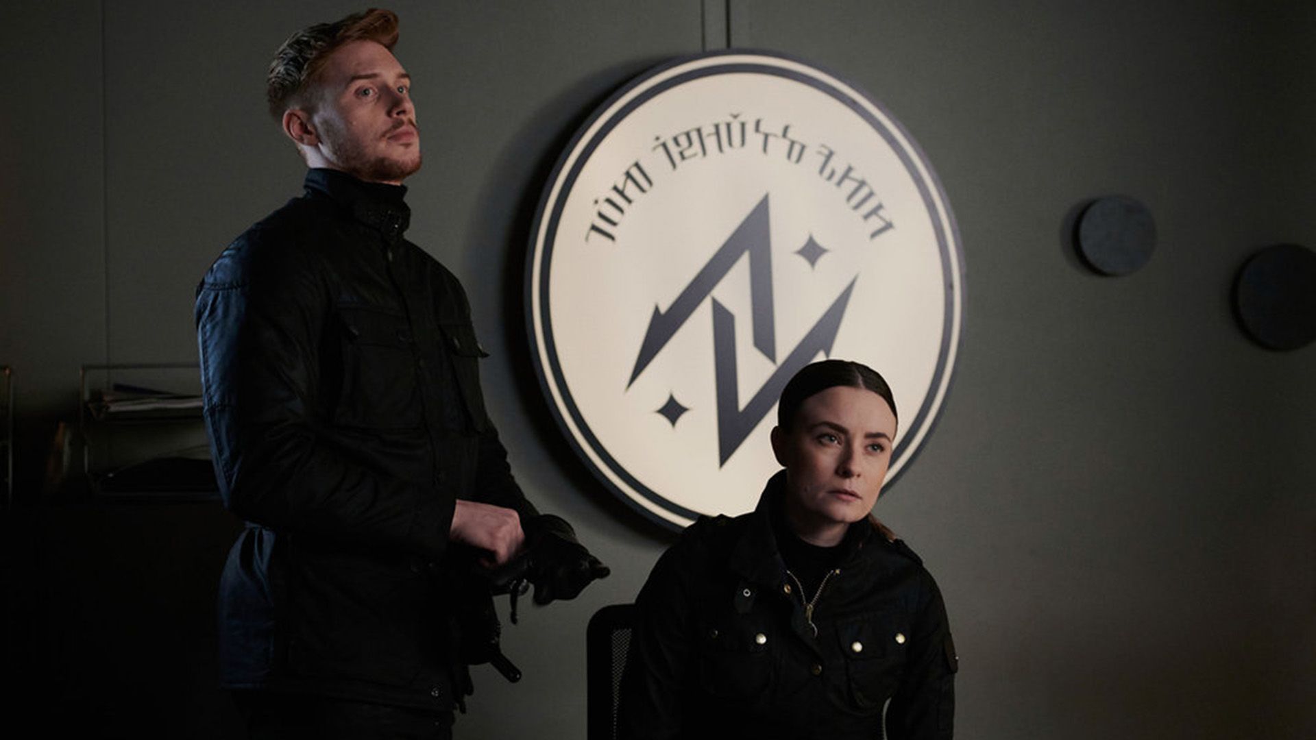 Max Parker in a black jacket stands against a glowing circular emblem backdrop with a woman seated at a table nearby
