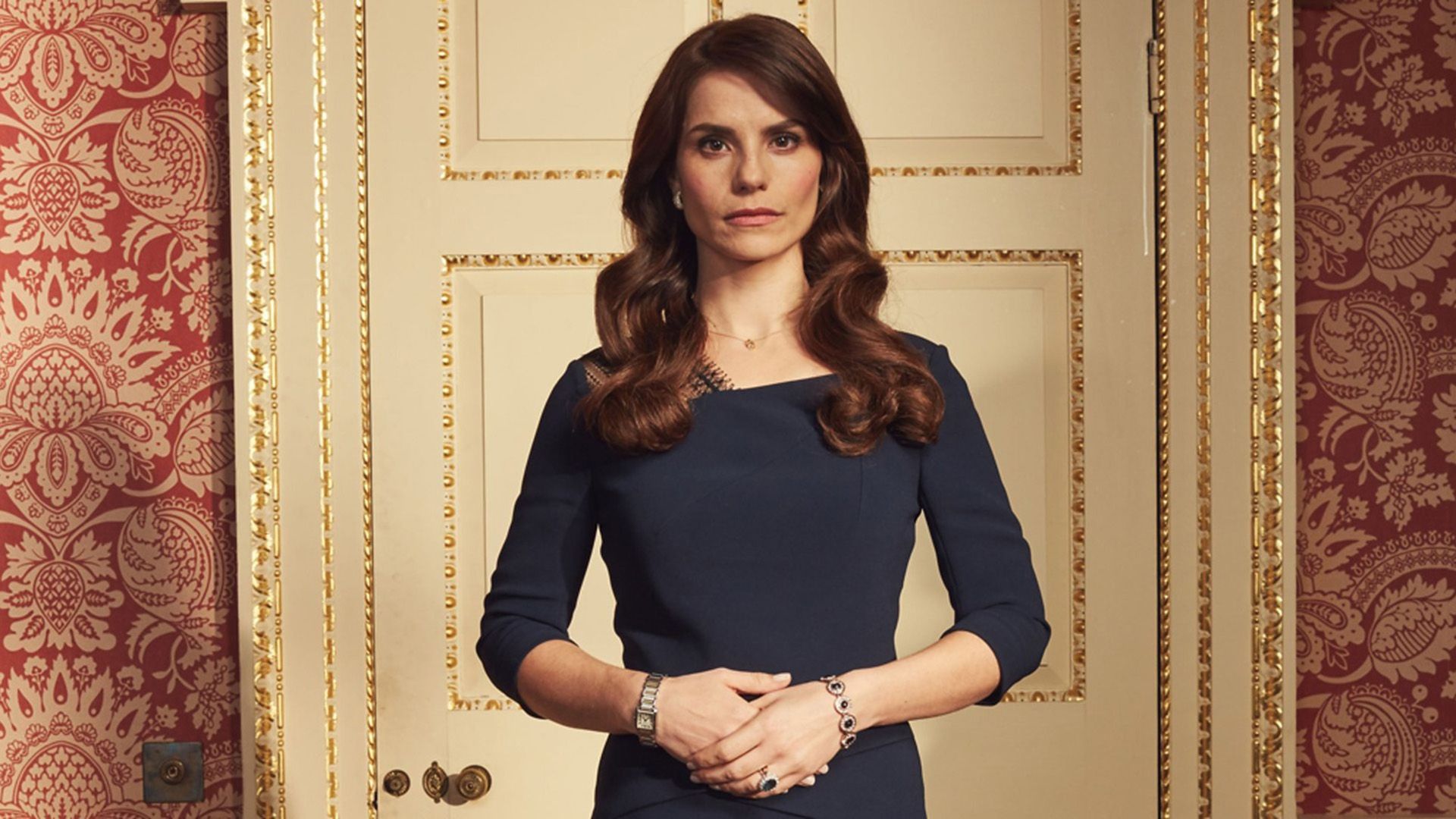 Charlotte Riley in a blue dress stands in front of a white door with gold trim