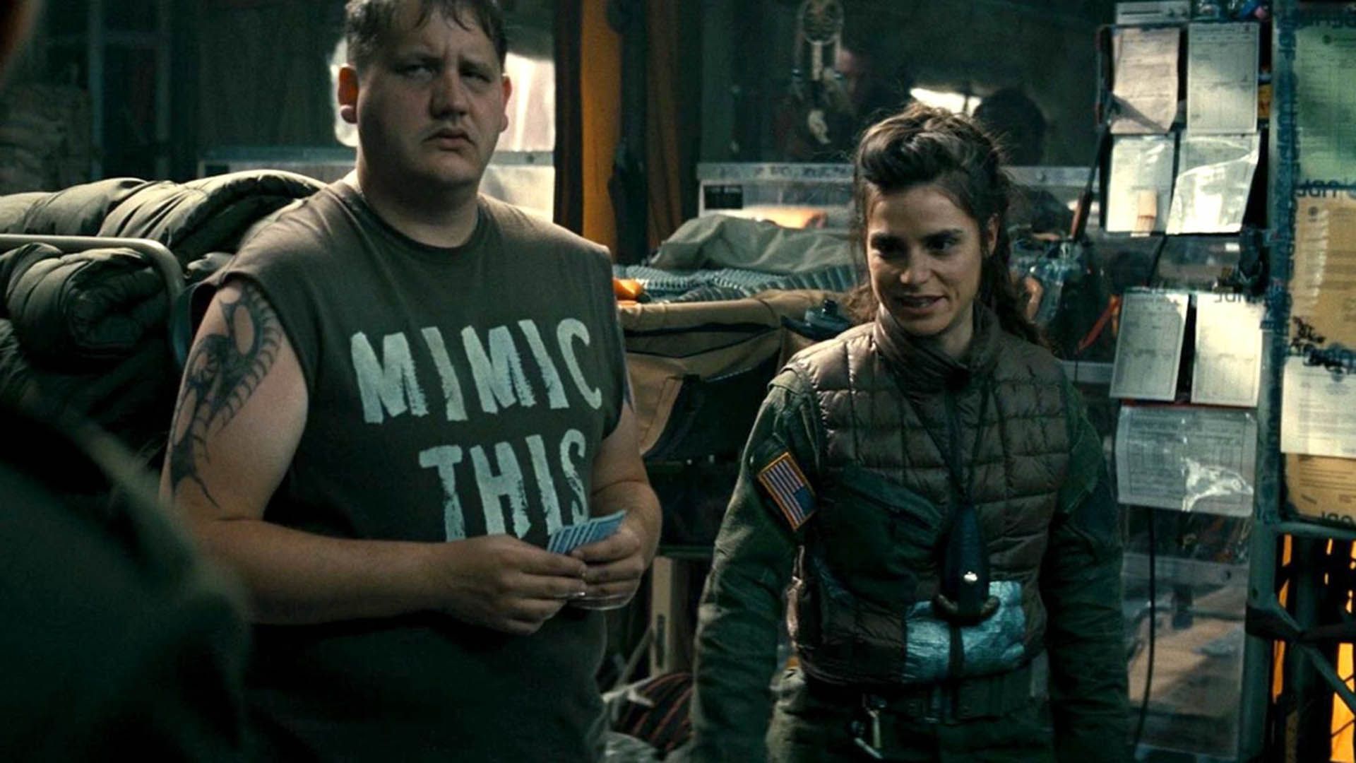 Charlotte Riley in khaki uniform stands next to a large man in a black tank top