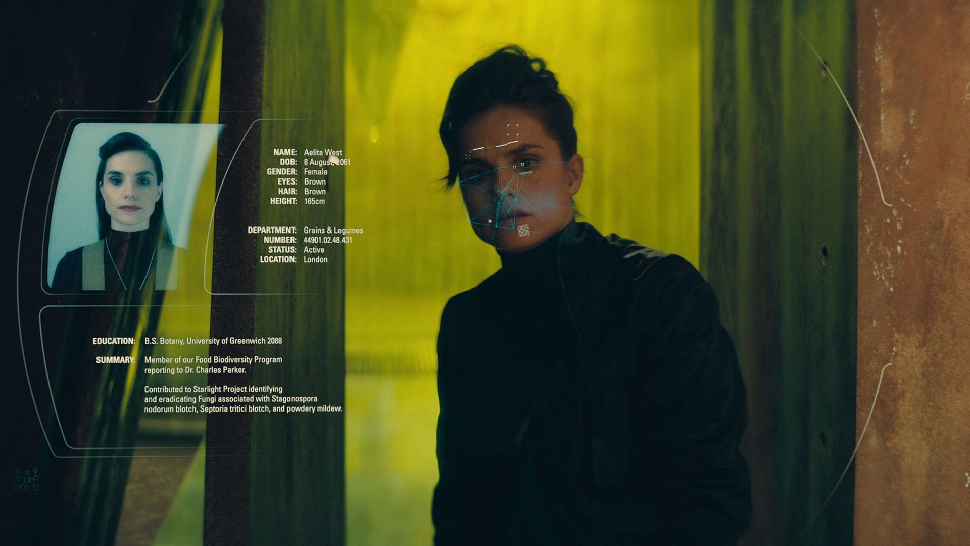 Charlotte Riley stands in a dark corridor with yellow-green lighting while a screen nearby displays her photograph and information about her