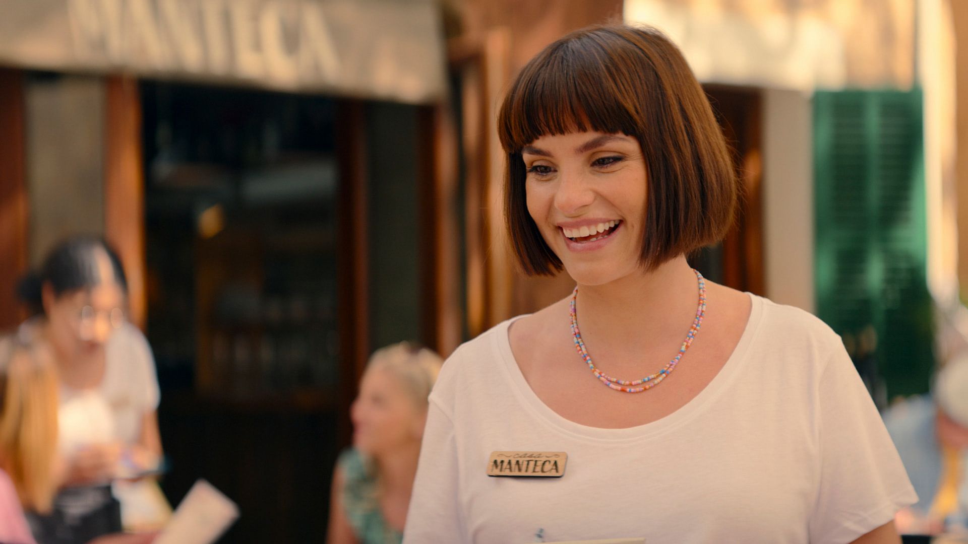 Charlotte Riley with a bob haircut wearing a white t-shirt, a wooden badge on her chest, and a colorful beaded necklace