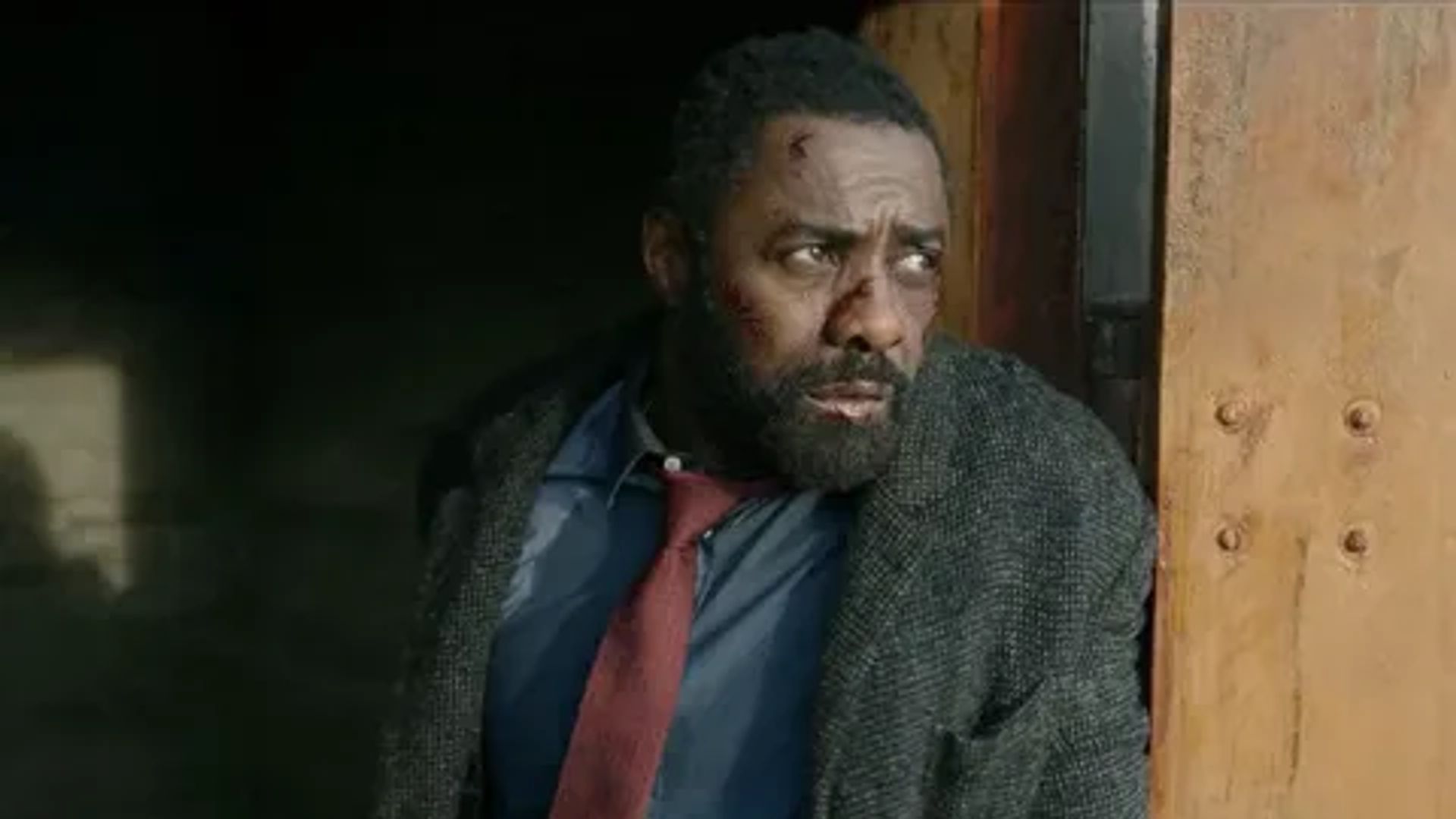 Idris Elba in a coat with a red tie looking to the side, film still