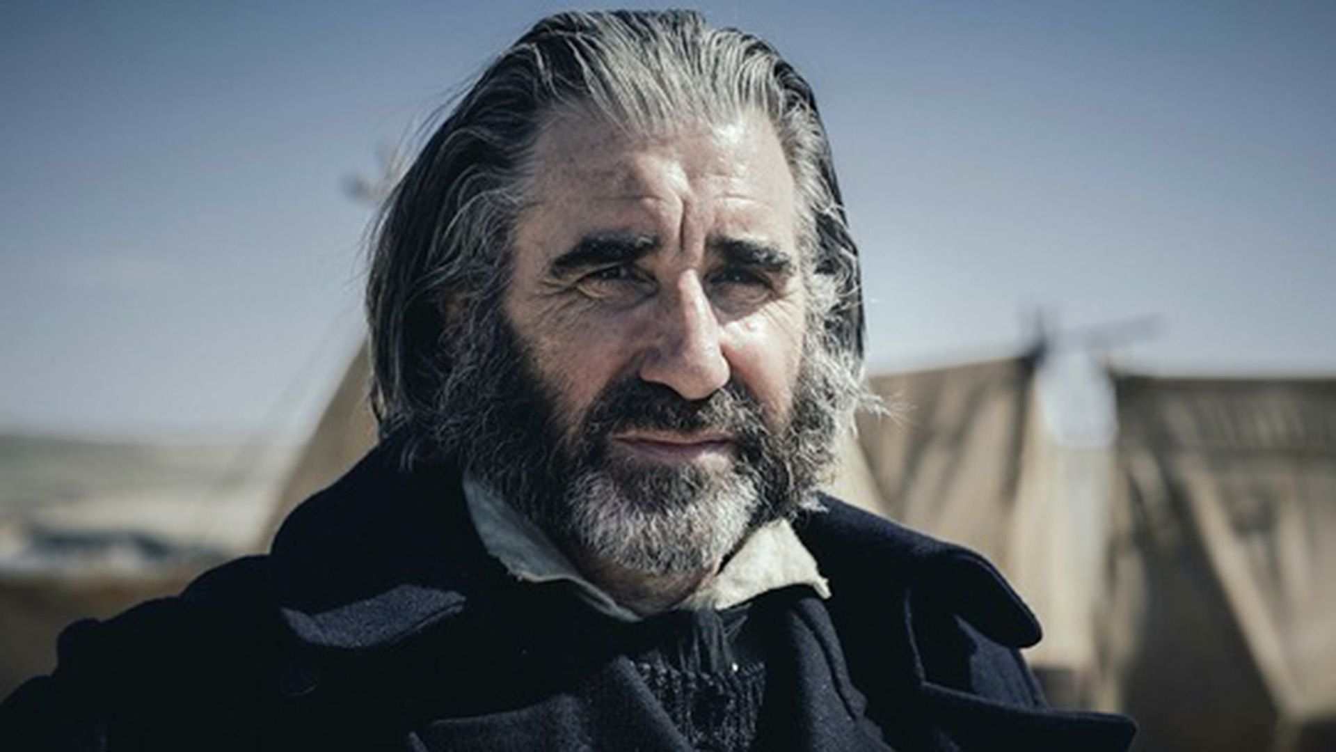 John Lynch with a gray beard squints in the sunlight