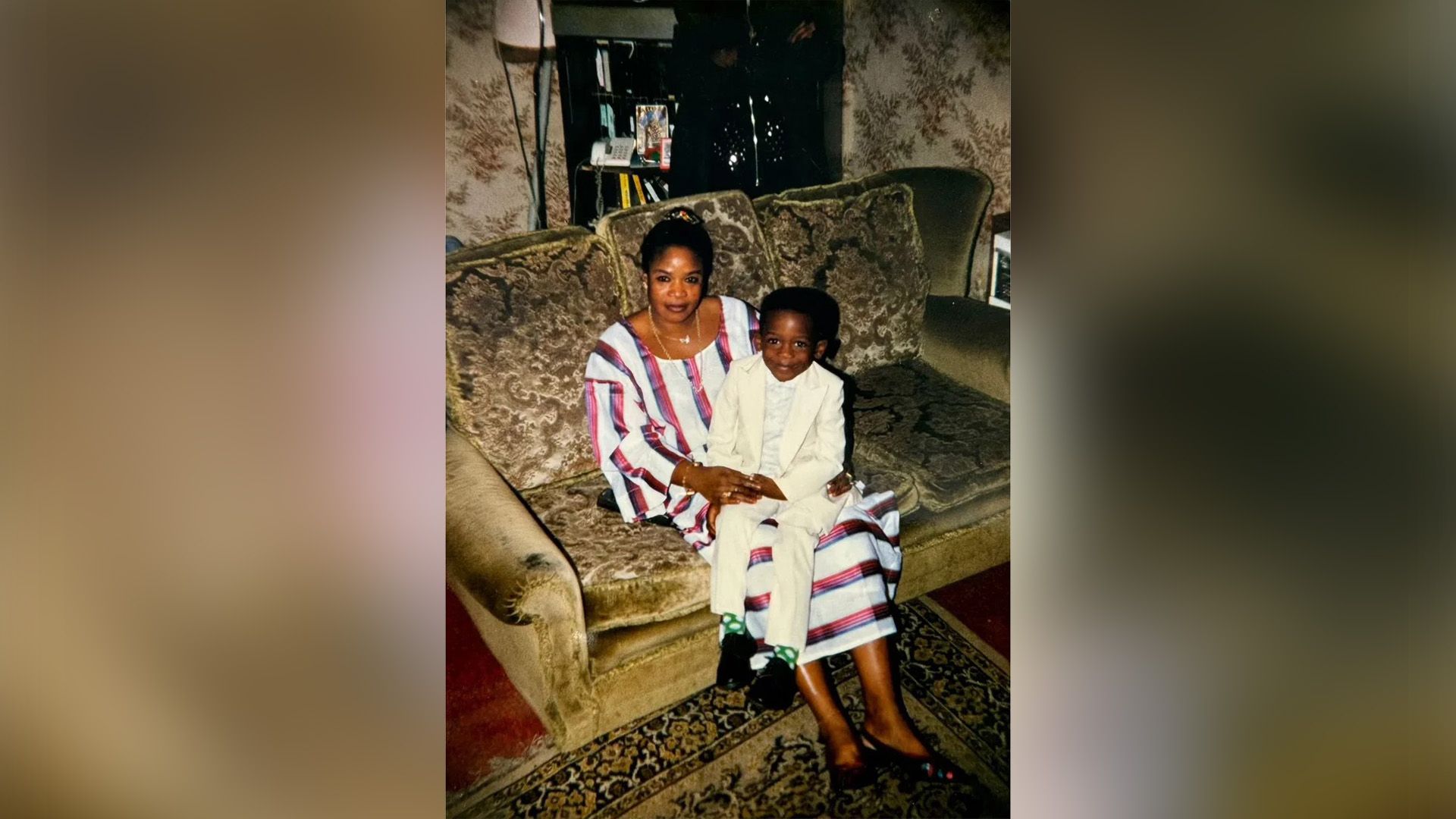 Little Damson Idris in a white suit sits on his mother's lap as she sits on the couch