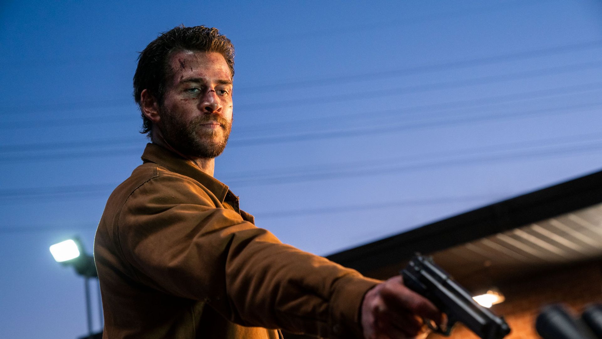 Liam Hemsworth in a brown jacket holding a gun