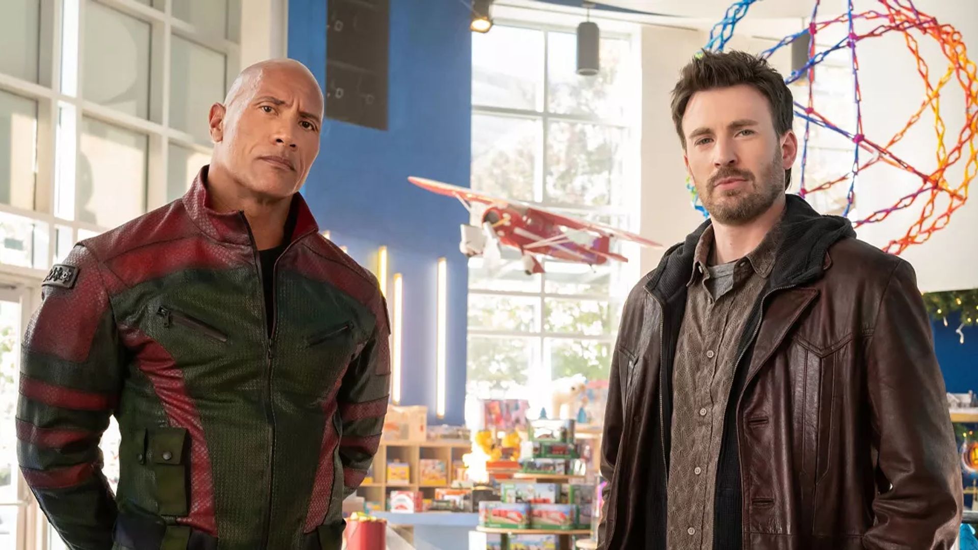 Chris Evans and Dwayne Johnson wearing jackets stand in a brightly lit room with toys and a model airplane in the background