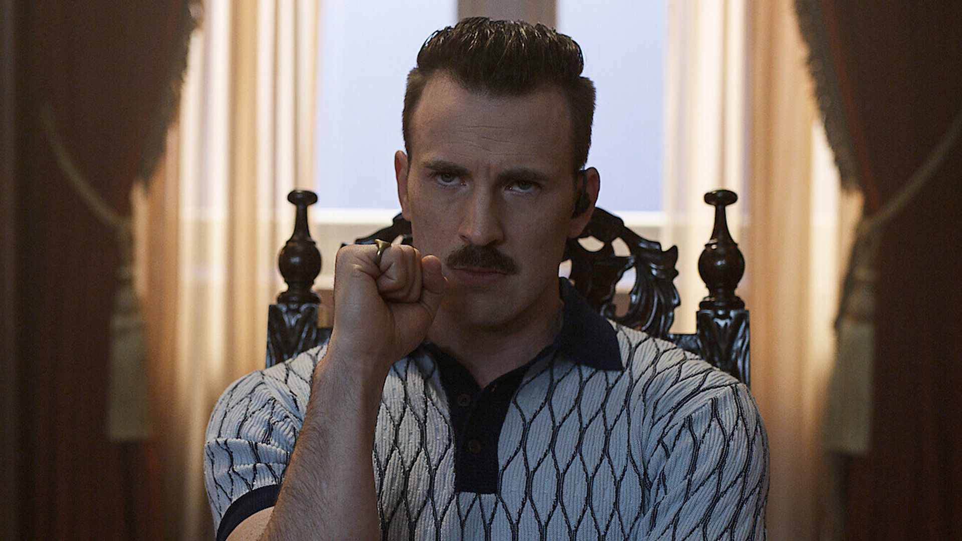 Chris Evans with a mustache wearing a patterned shirt sits in an ornate chair gazing pensively ahead with his fist clenched against his face