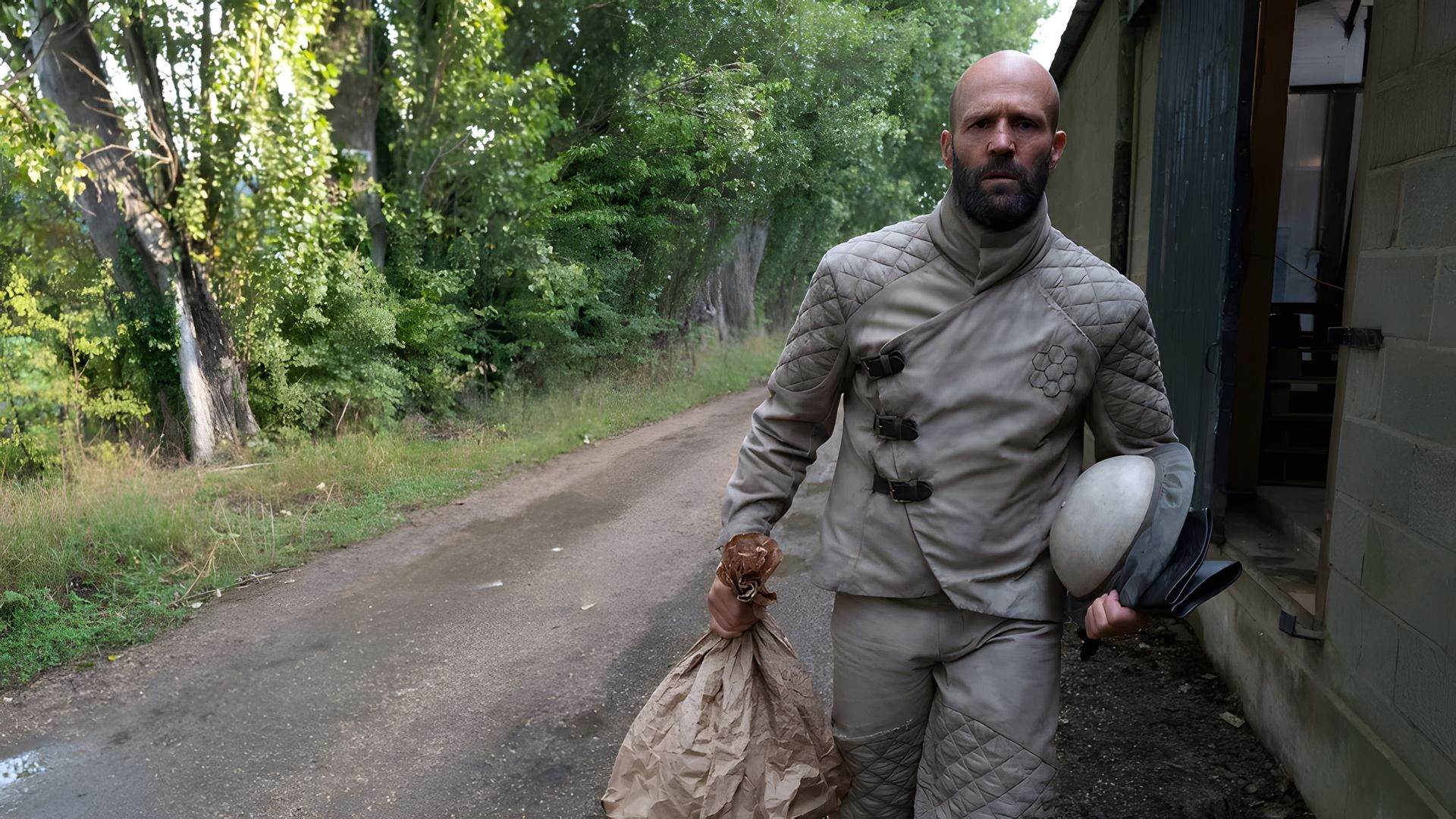 Jason Statham in a gray protective suit walking down a road carrying a bag and helmet with trees in the background