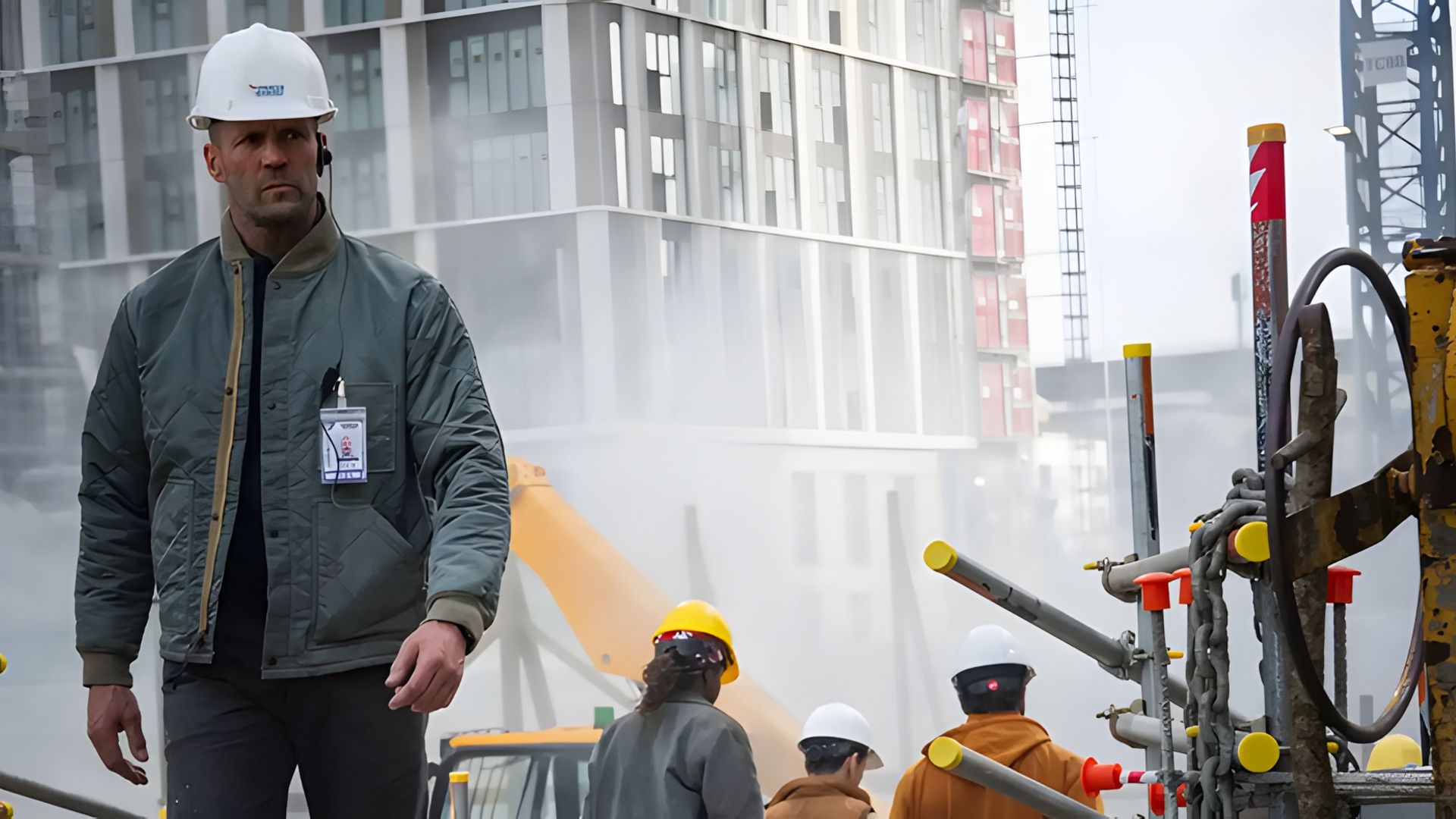 Jason Statham in a hard hat and jacket walks through a construction site with workers and machinery visible in the hazy background