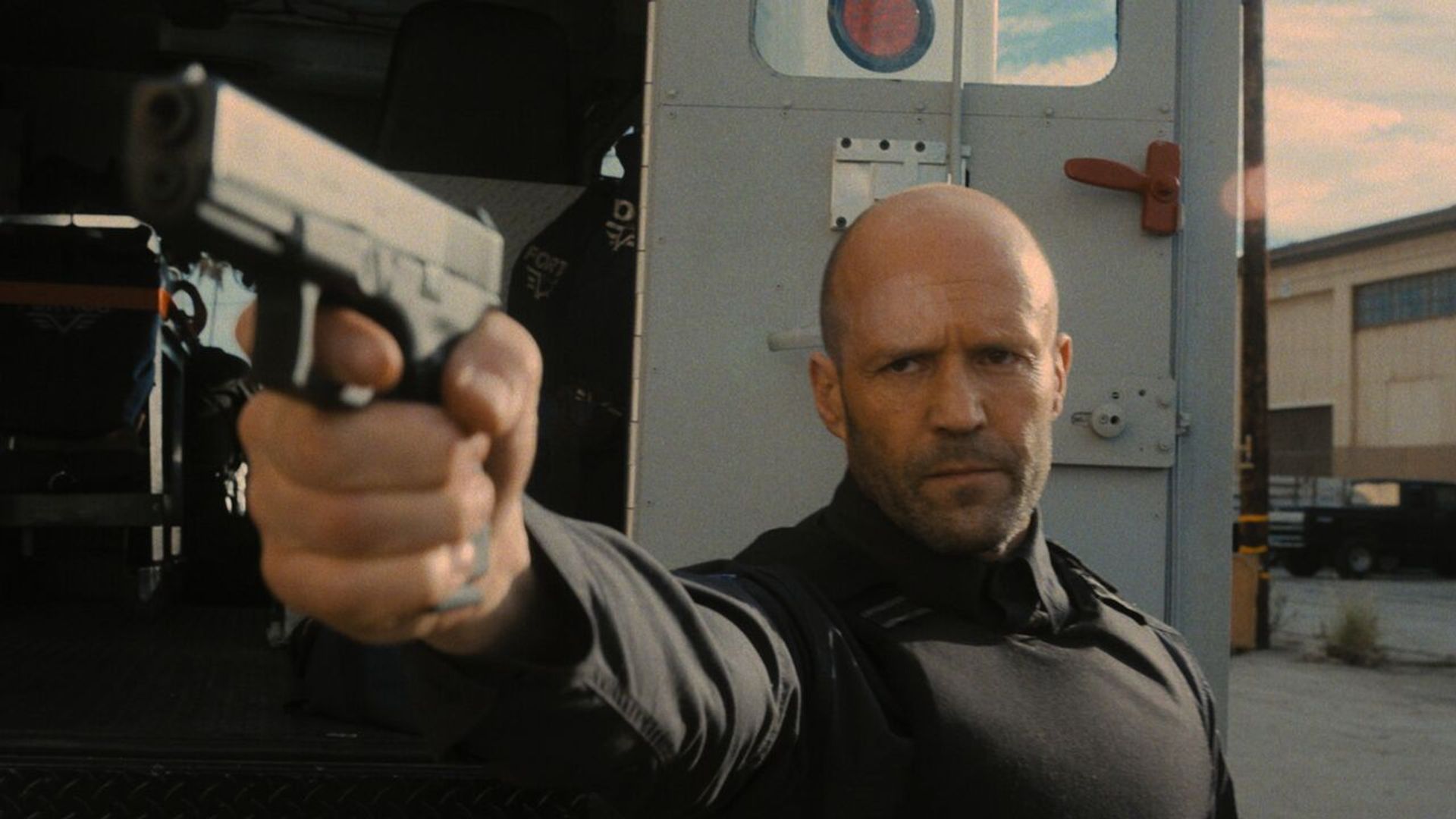 Jason Statham in black tactical gear aims a pistol while standing by an armored van against an industrial backdrop