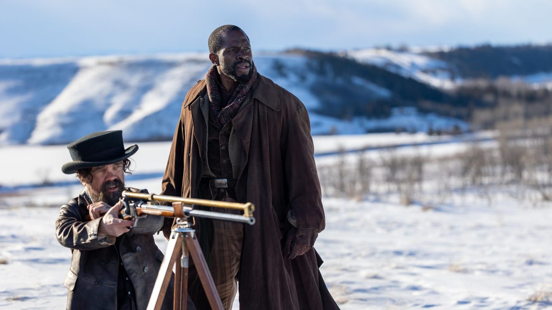 Peter Dinklage and Gbenga Akinnagbe stand in the snow, one aiming a rifle mounted on a tripod, the other gazing into the distance