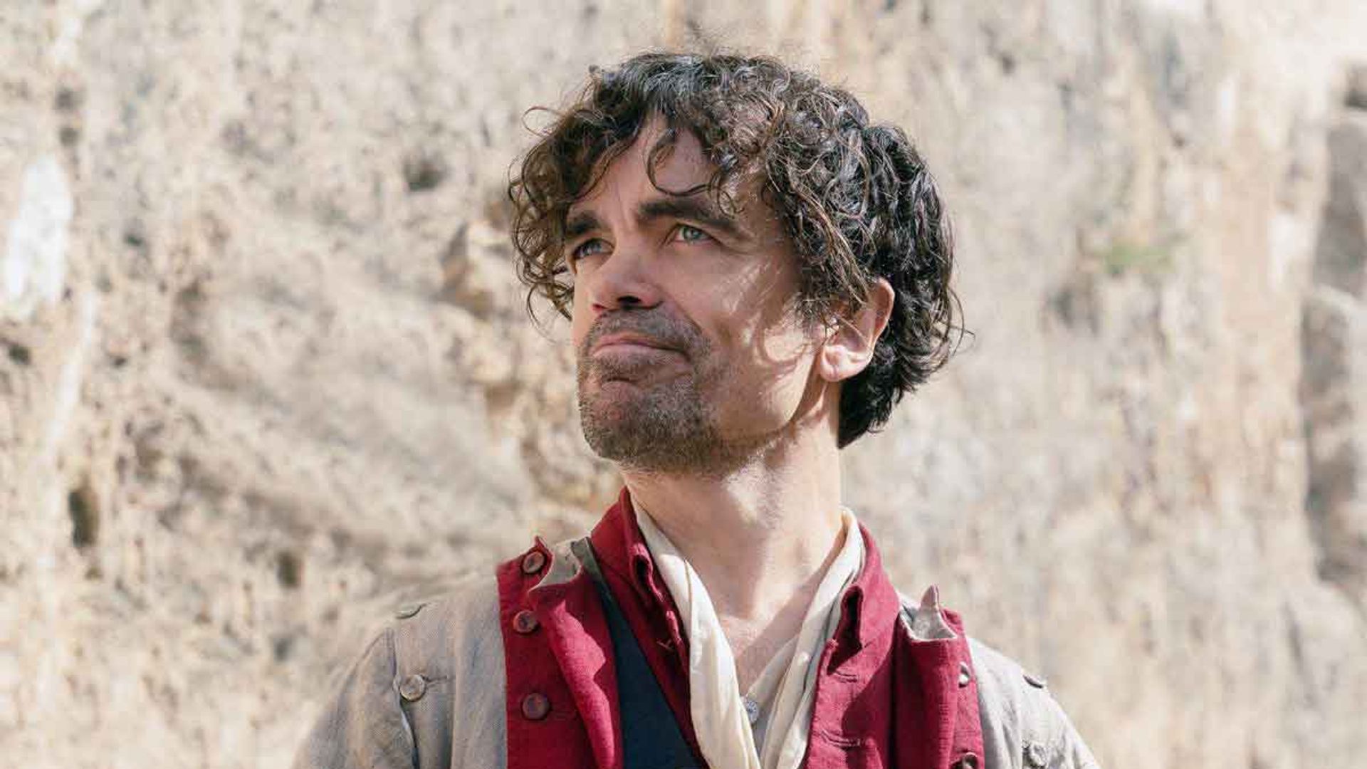 Peter Dinklage in period costume with a red doublet holds a letter and looks up with a slight smile