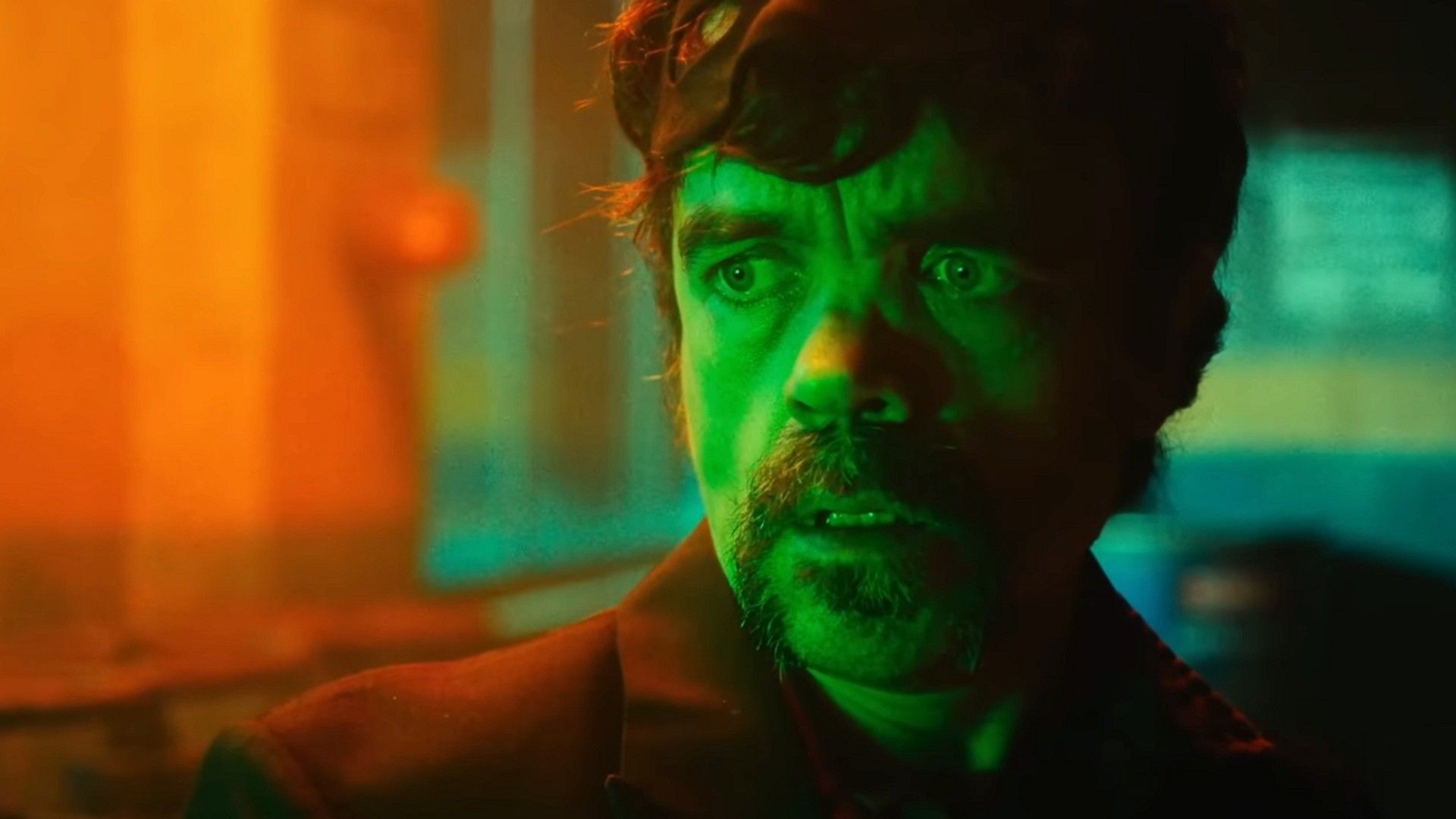 Peter Dinklage stands in a dark room illuminated by green and orange light