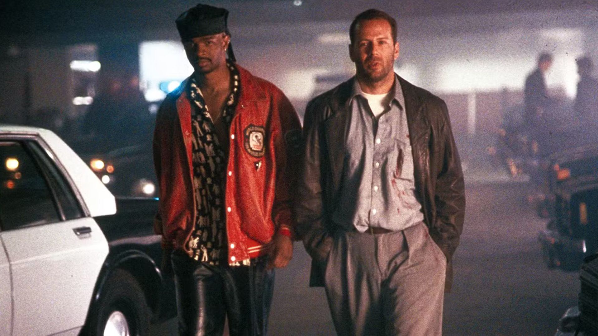 A young black man in a red sports jacket and a young white man in a leather jacket are walking through an underground parking lot