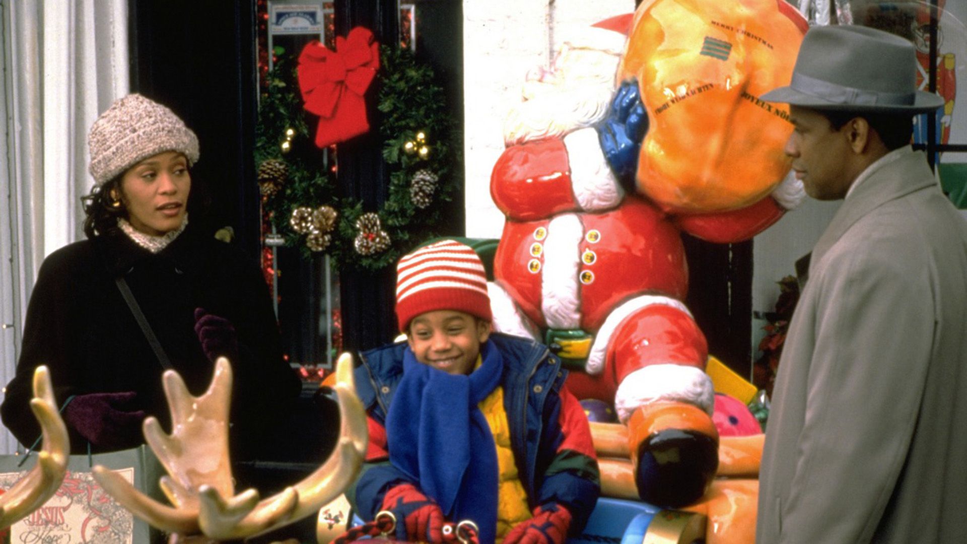 A dark-skinned family - mother, father, and son, dressed in outerwear, talking to each other next to a large figure of Santa Claus