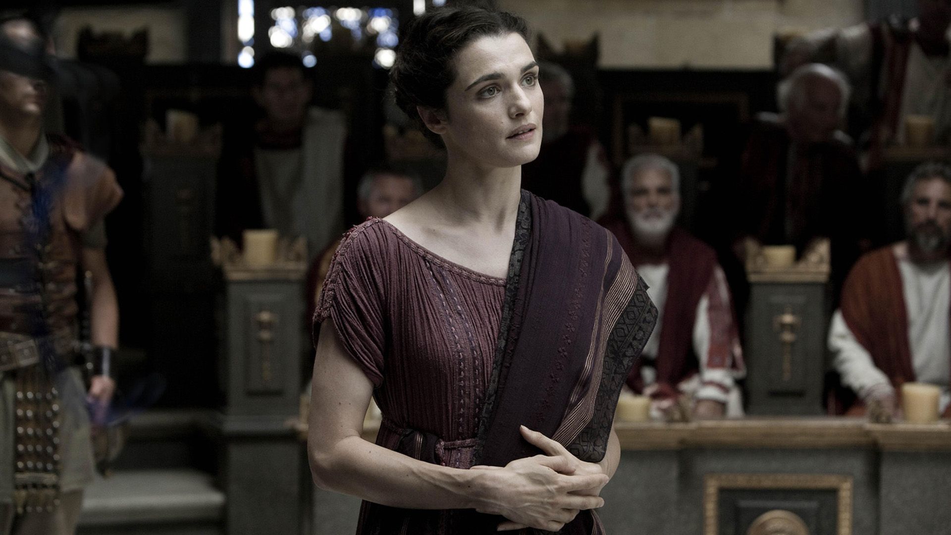 A young dark-haired woman in burgundy ancient clothing, against the background of an older man in ancient attire