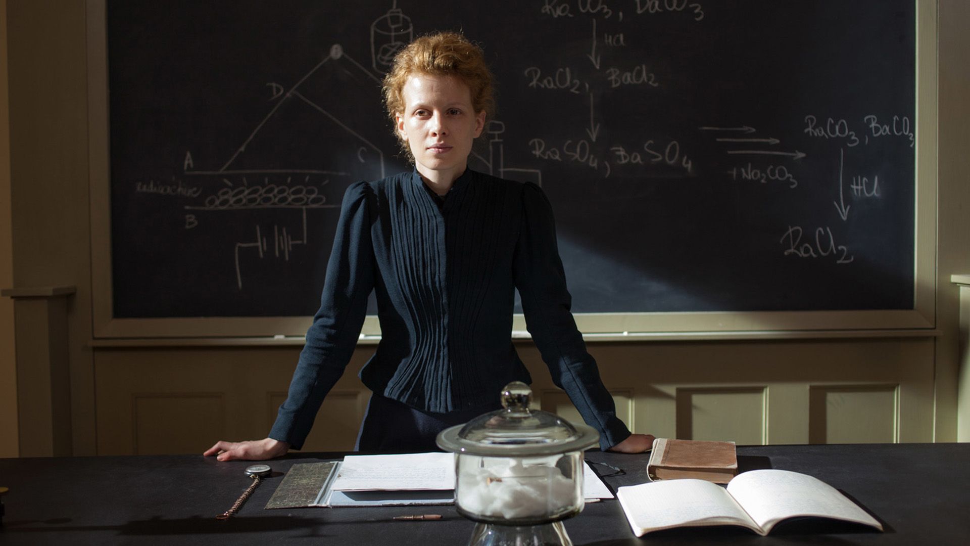 A young red-haired woman in a strict black dress stands at a table by a chalkboard with formulas written in chalk