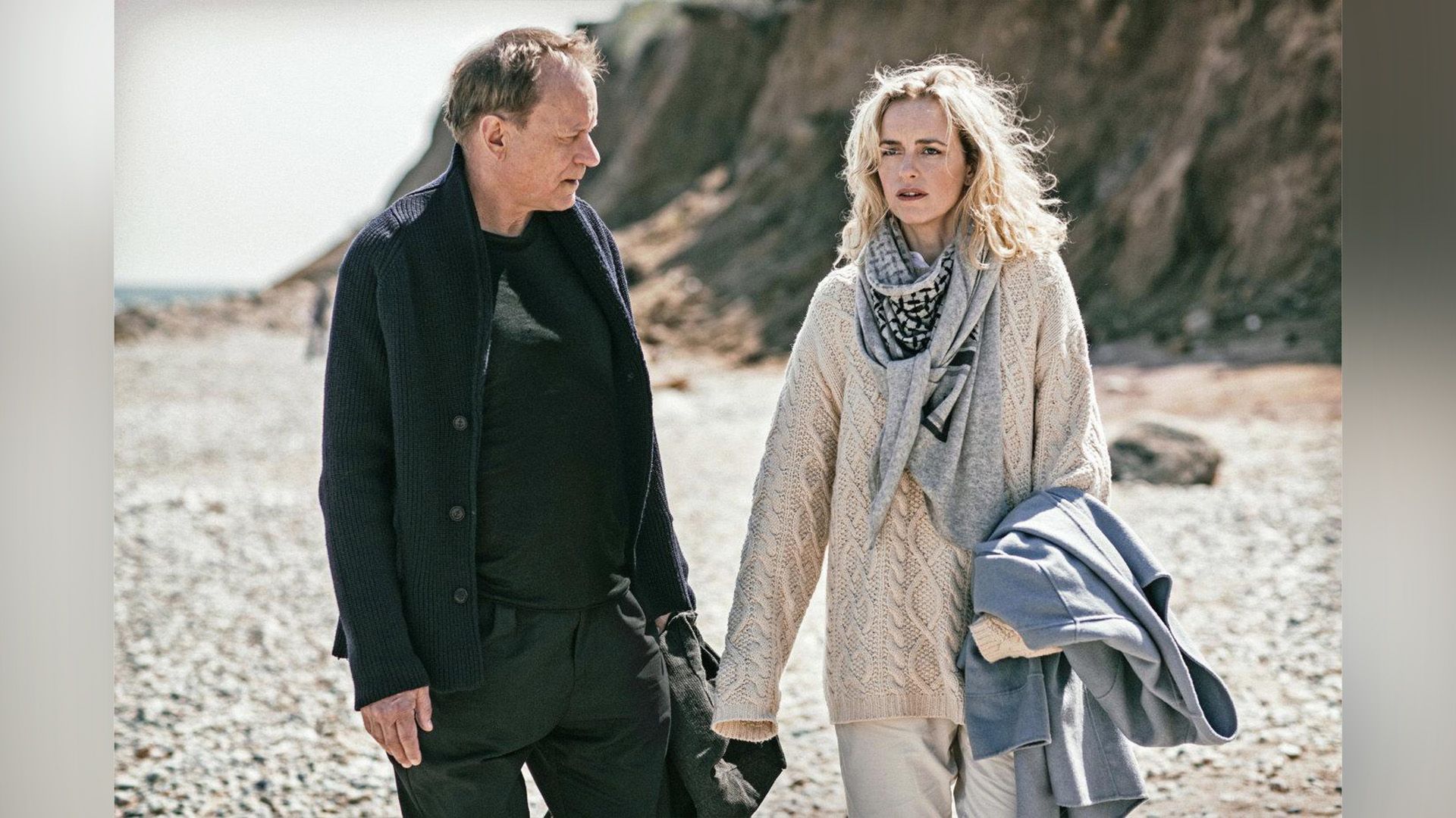 A man and a light-haired woman, both of age, are standing on the beach by a cliff