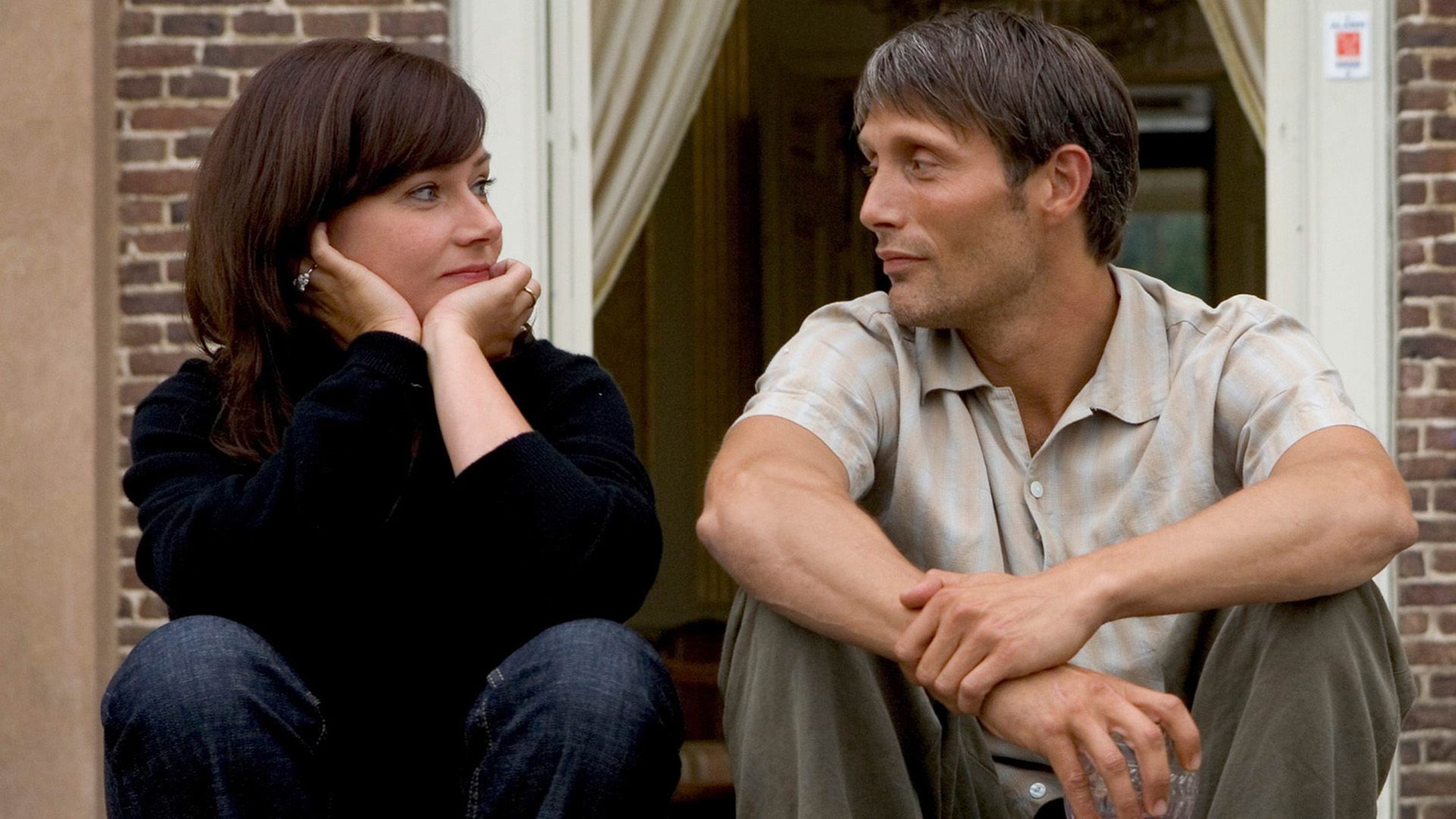 A man and a woman are sitting on the porch of a brick house by the open door, looking at each other