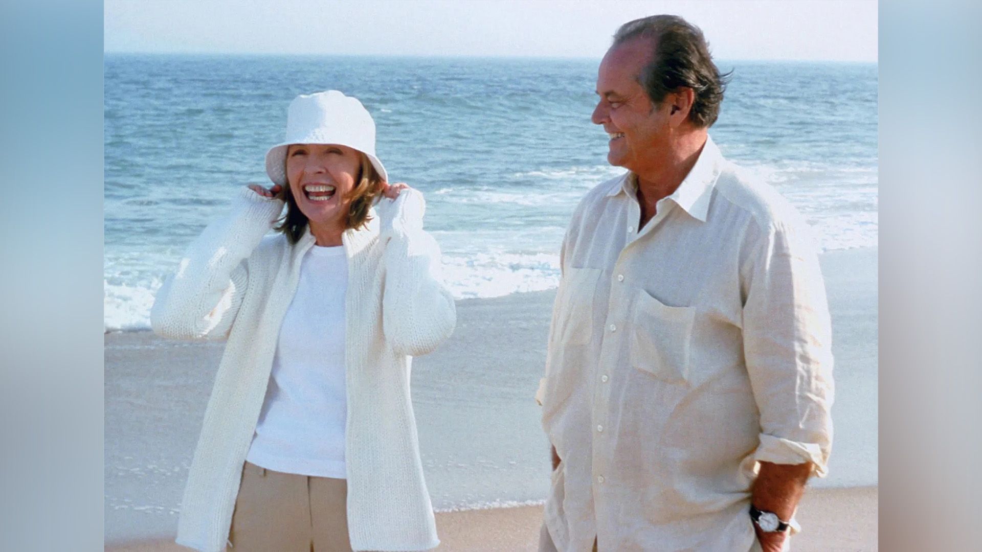 An elderly man and woman, both dressed in white, are standing on the beach by the water, the woman is laughing