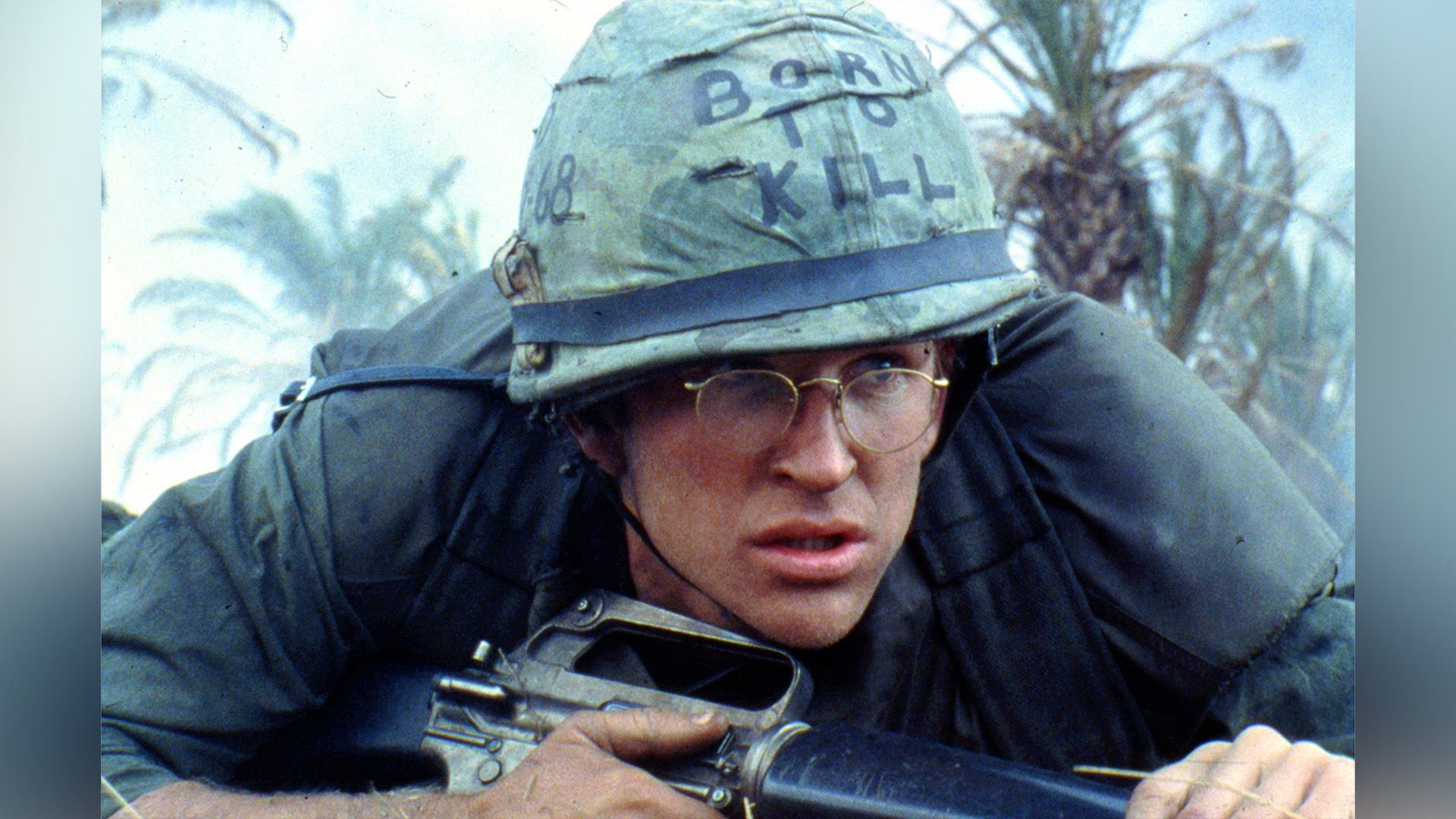 A thin young man in glasses and a military helmet lies with a weapon in his hands against a backdrop of palm trees