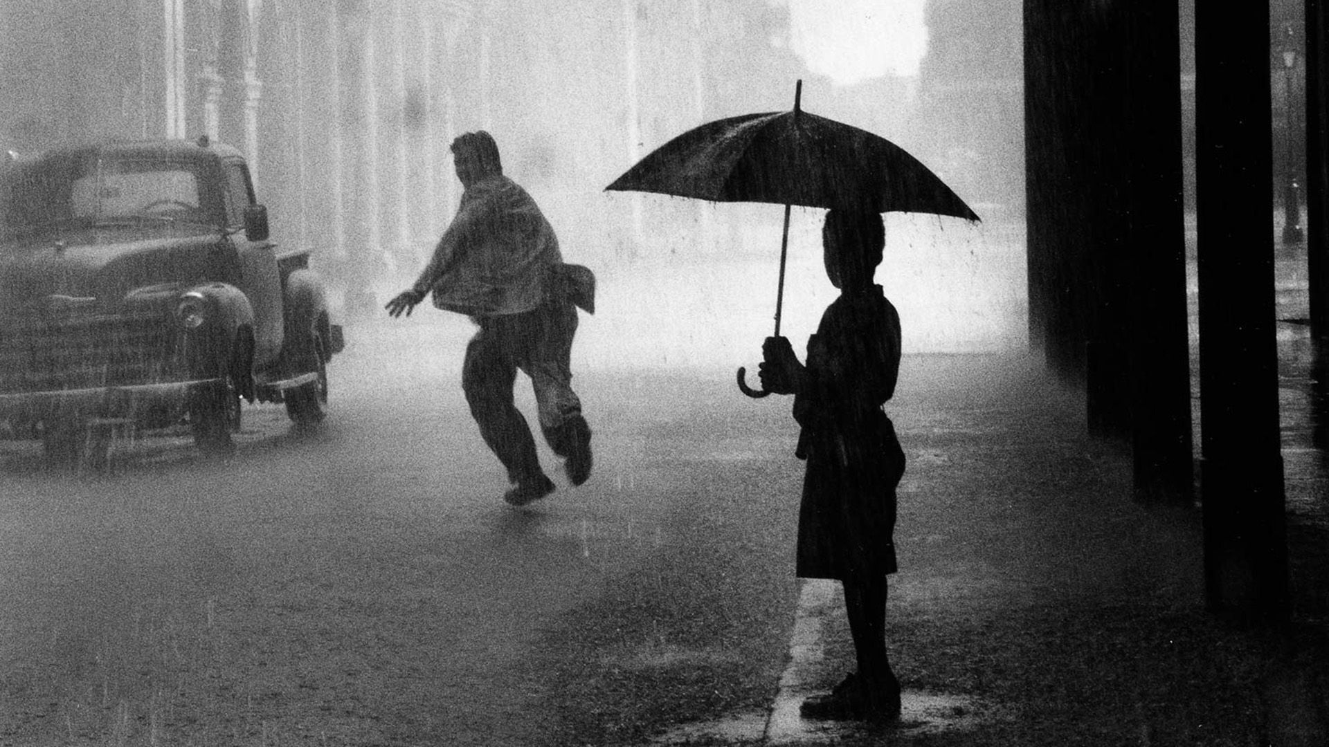 In the foreground, a silhouette of a boy under an umbrella, watching a man running away in the rain along the wall