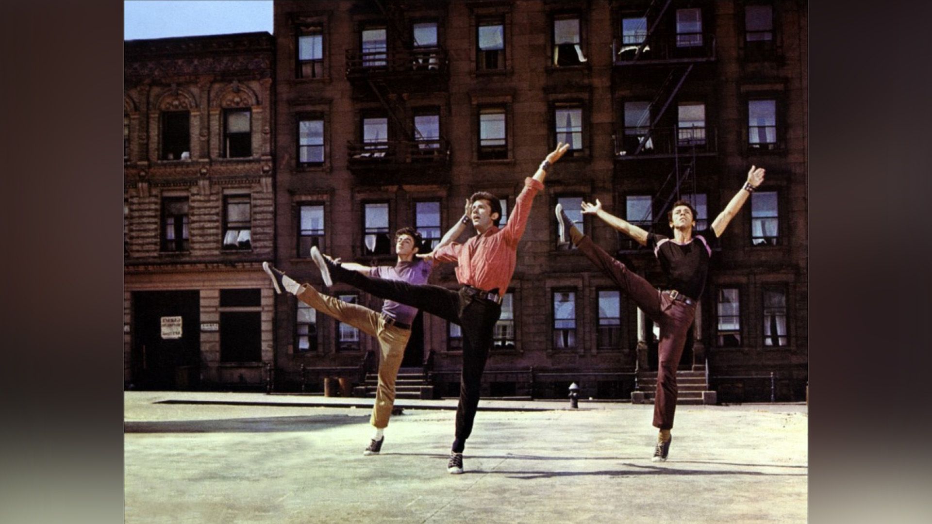 The courtyard in front of the apartment building, three young men in jeans and t-shirts of different colors are dancing